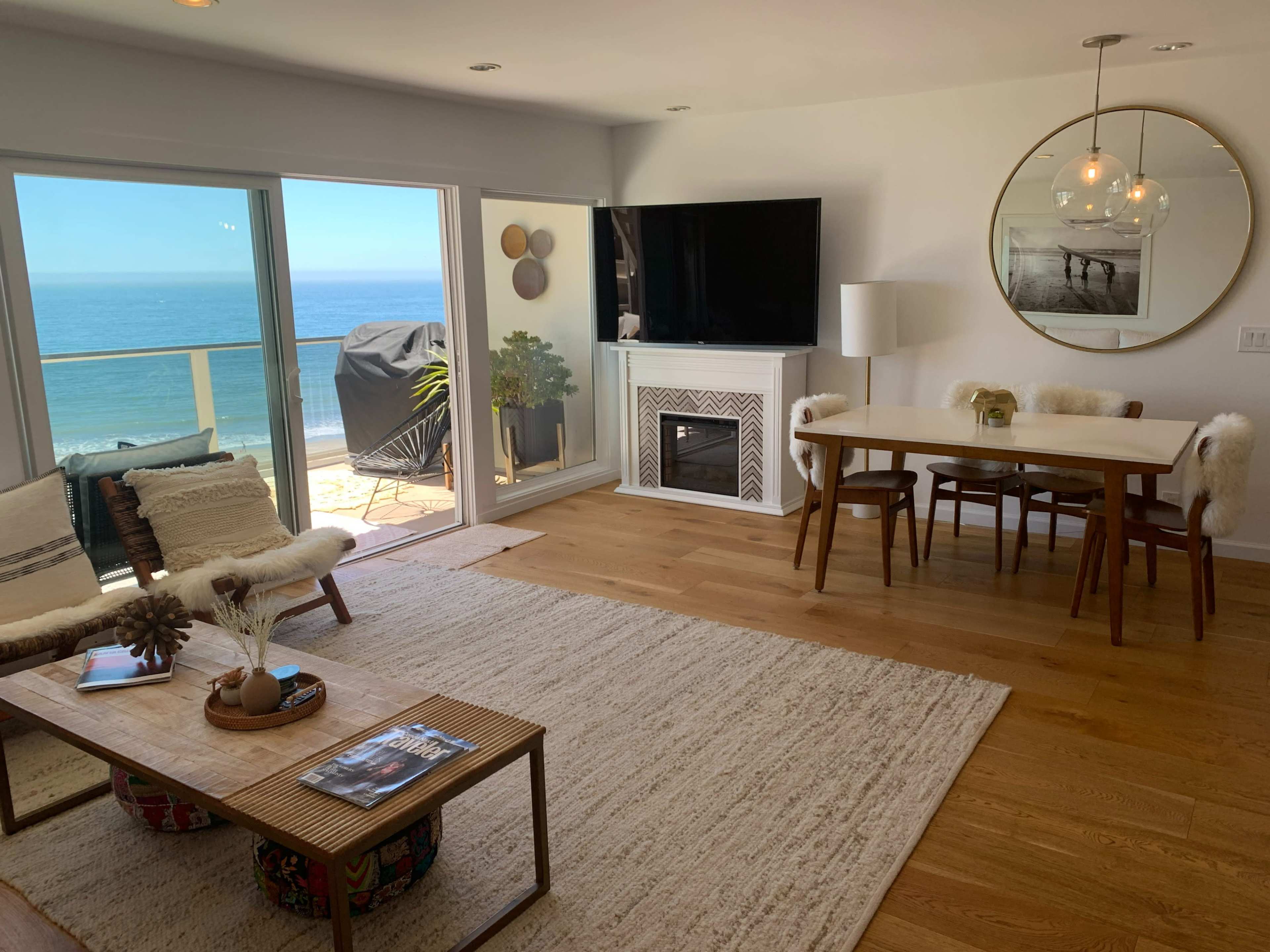 A modern living room with a dining table, a fireplace, and large windows overlooking the ocean.