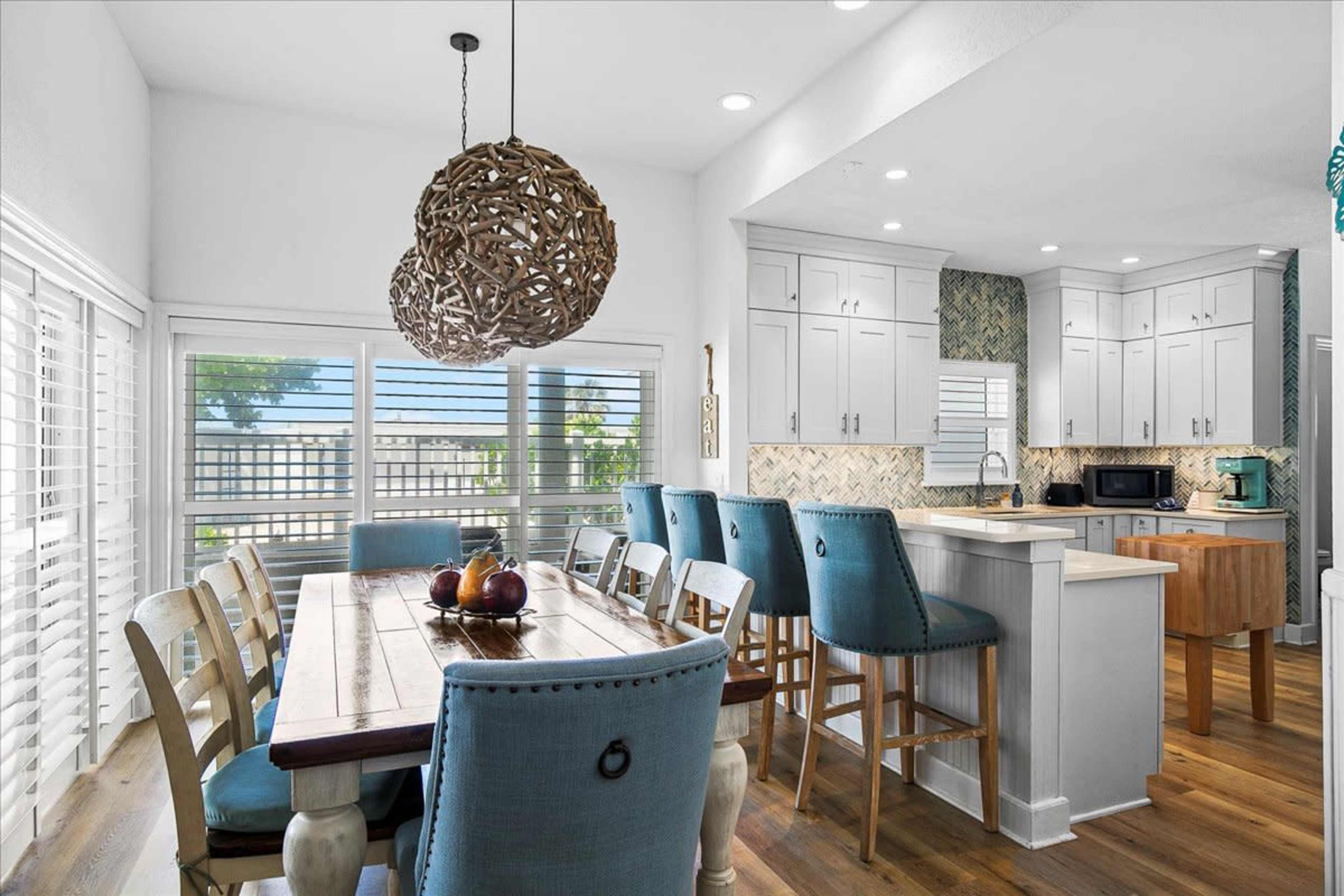 The image shows a modern kitchen and dining area featuring a large wooden dining table surrounded by chairs, with a pendant light above and the kitchen visible in the background.