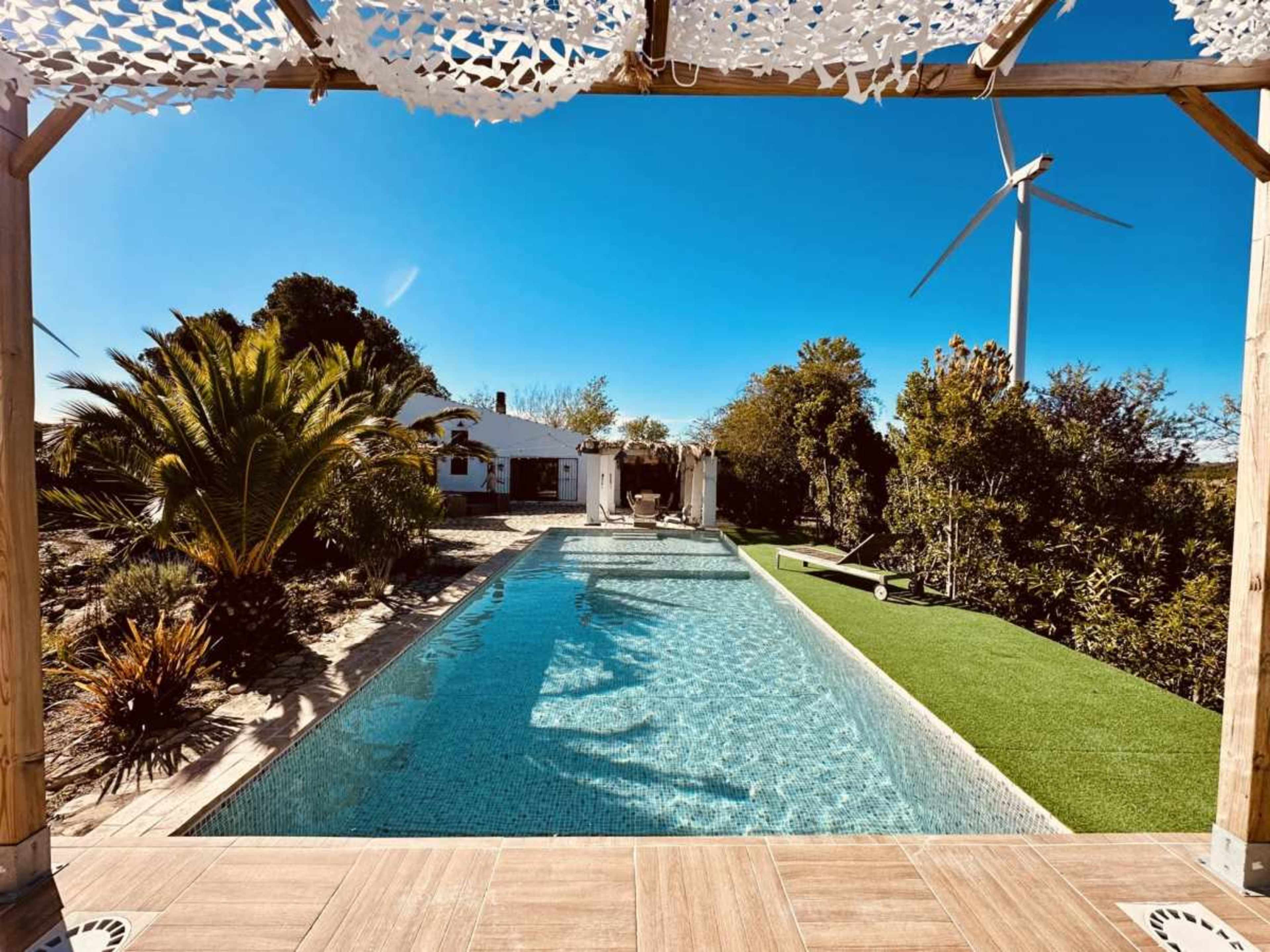 The image shows a modern swimming pool surrounded by tropical plants and a wind turbine in the background, with a clear blue sky overhead.