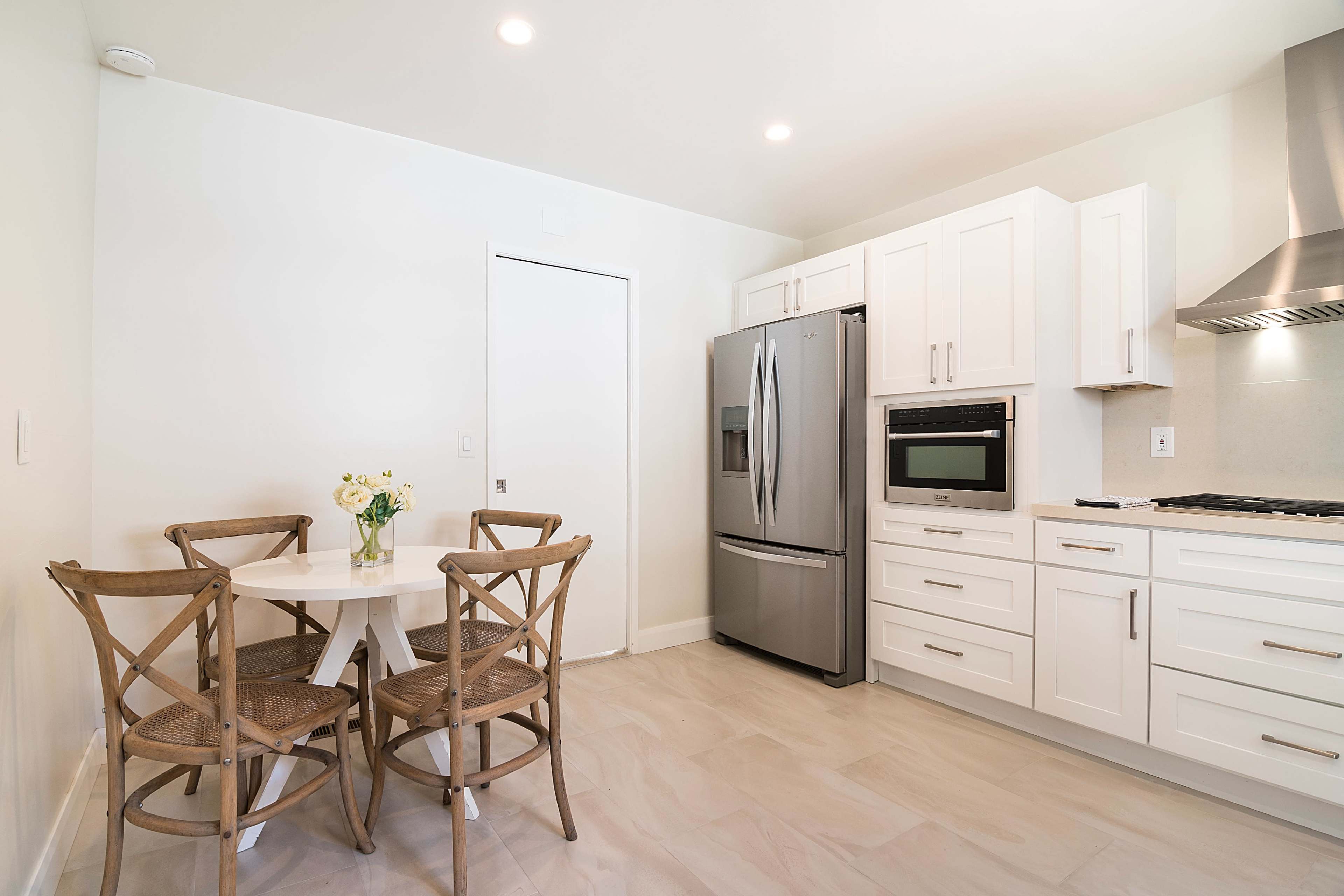 The image shows a modern kitchen featuring white cabinetry, stainless steel appliances, and a small dining table with four wooden chairs.