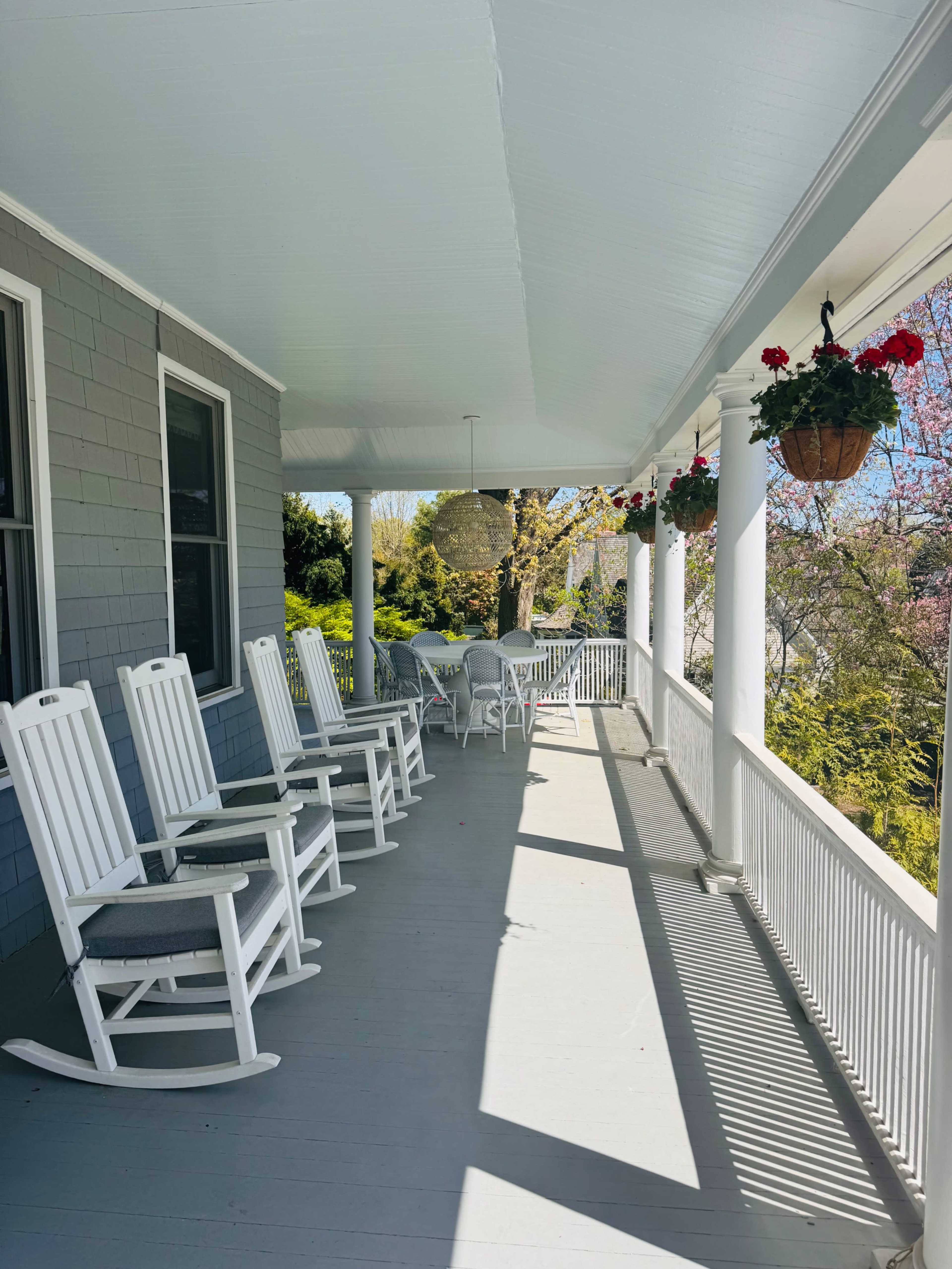 The image shows a porch with white rocking chairs lined up against a gray wall, adorned with hanging flower pots.