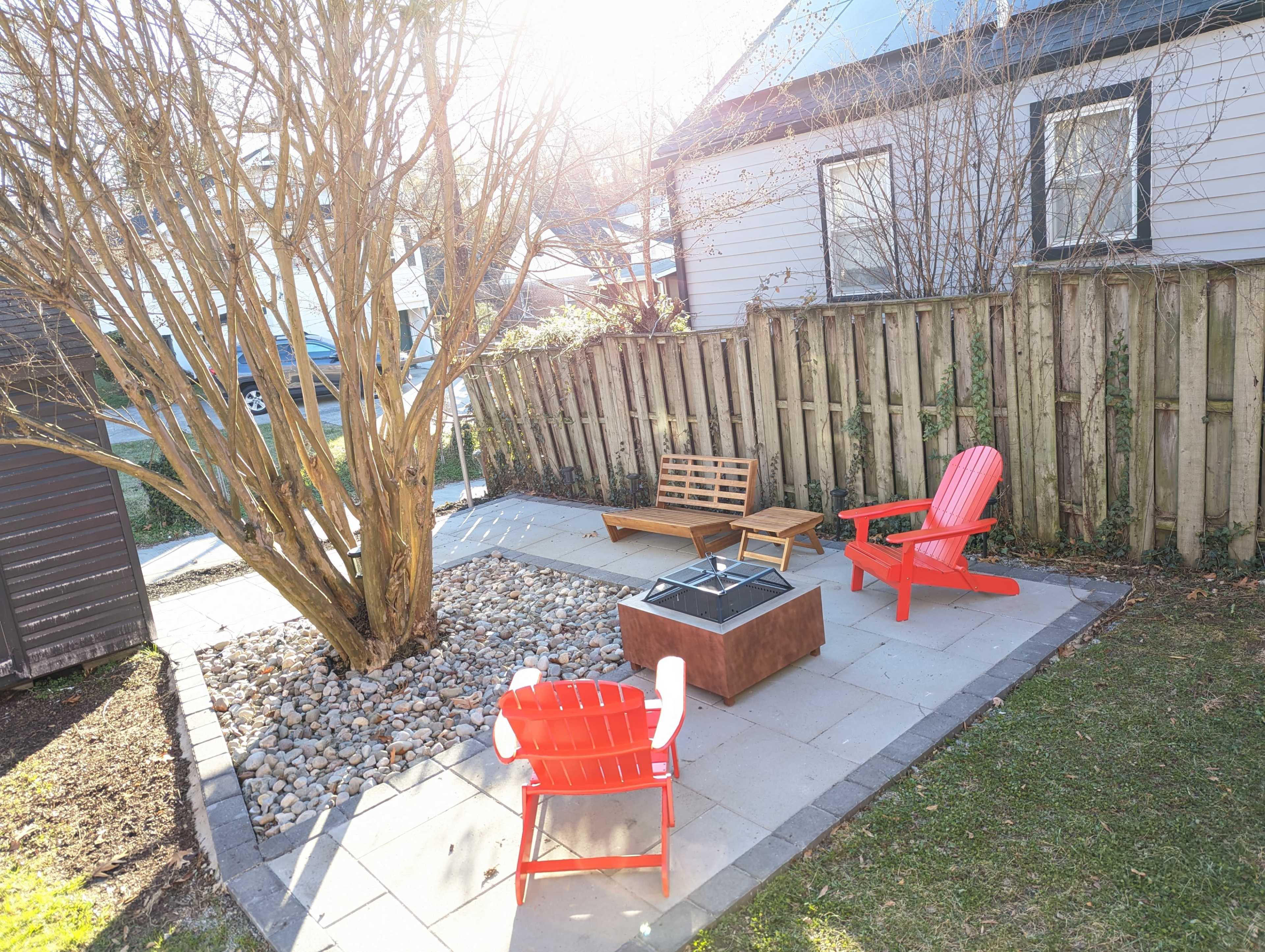 A small outdoor patio area features a stone fire pit surrounded by four colorful chairs and a tree, enclosed by a wooden fence.