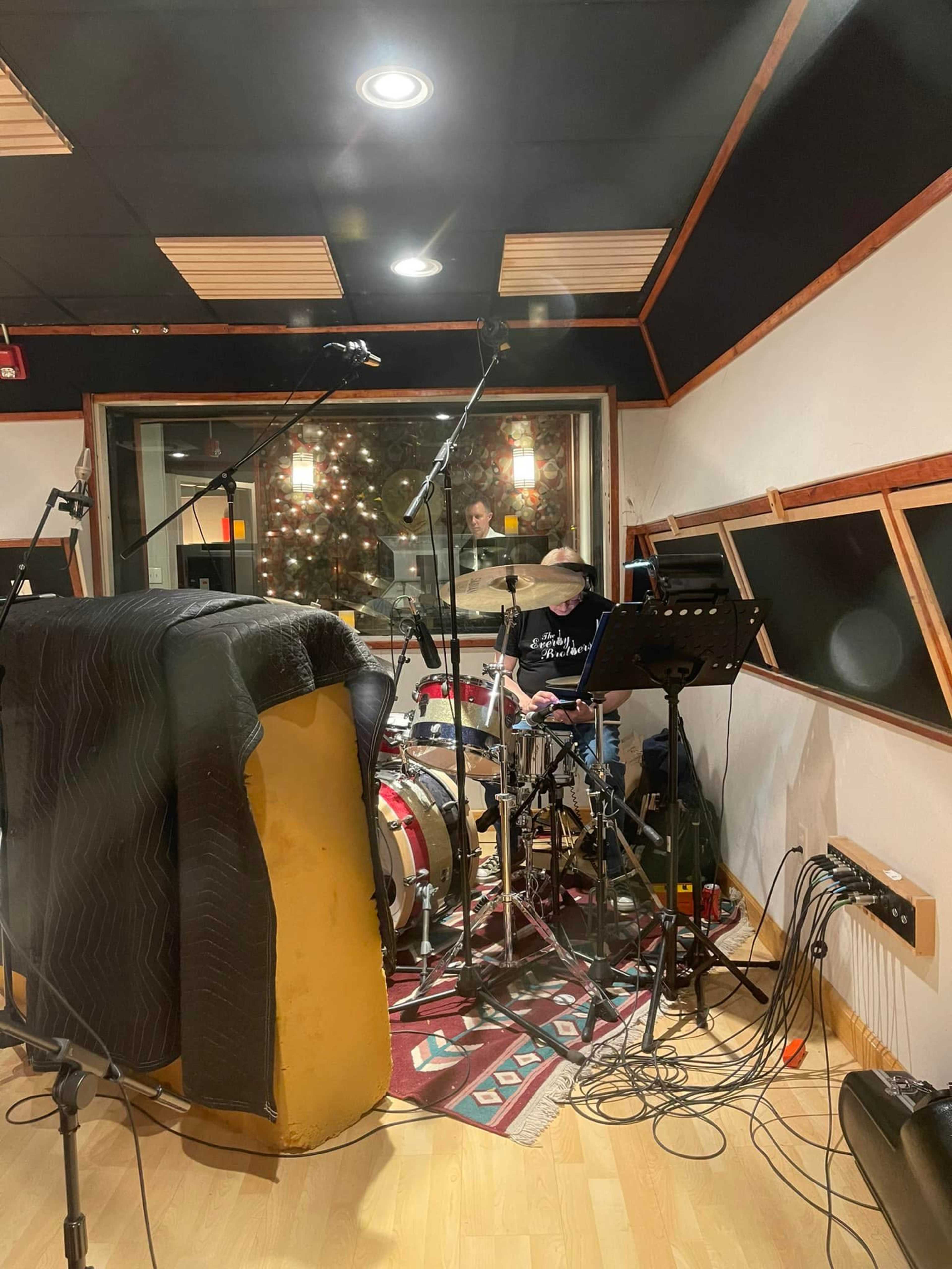 A drummer plays in a recording studio, surrounded by musical equipment and soundproofing materials.