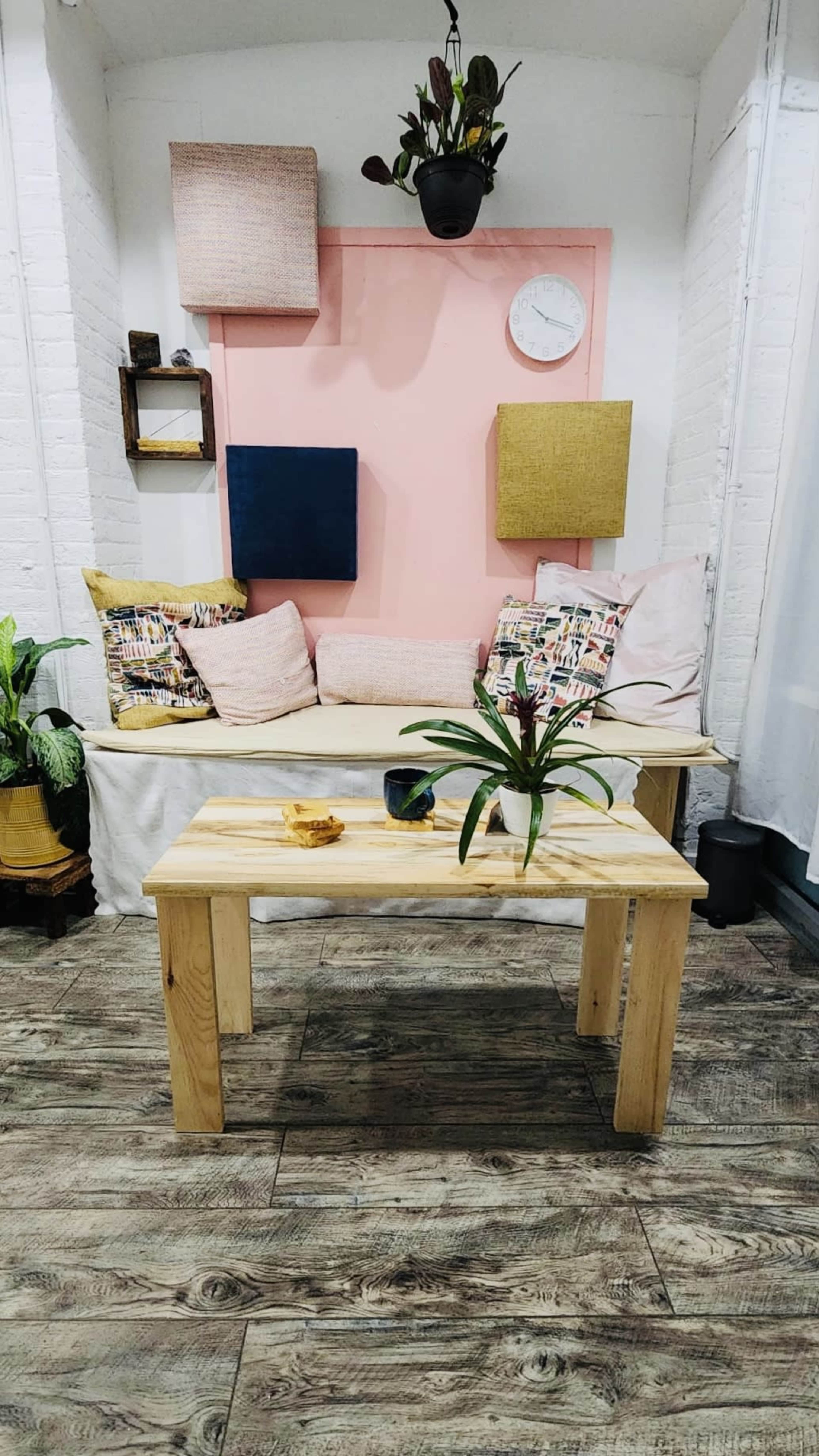 A cozy seating area with a wooden bench, decorative pillows, a coffee table with a plant, and various wall-mounted shelves against a pastel pink backdrop.