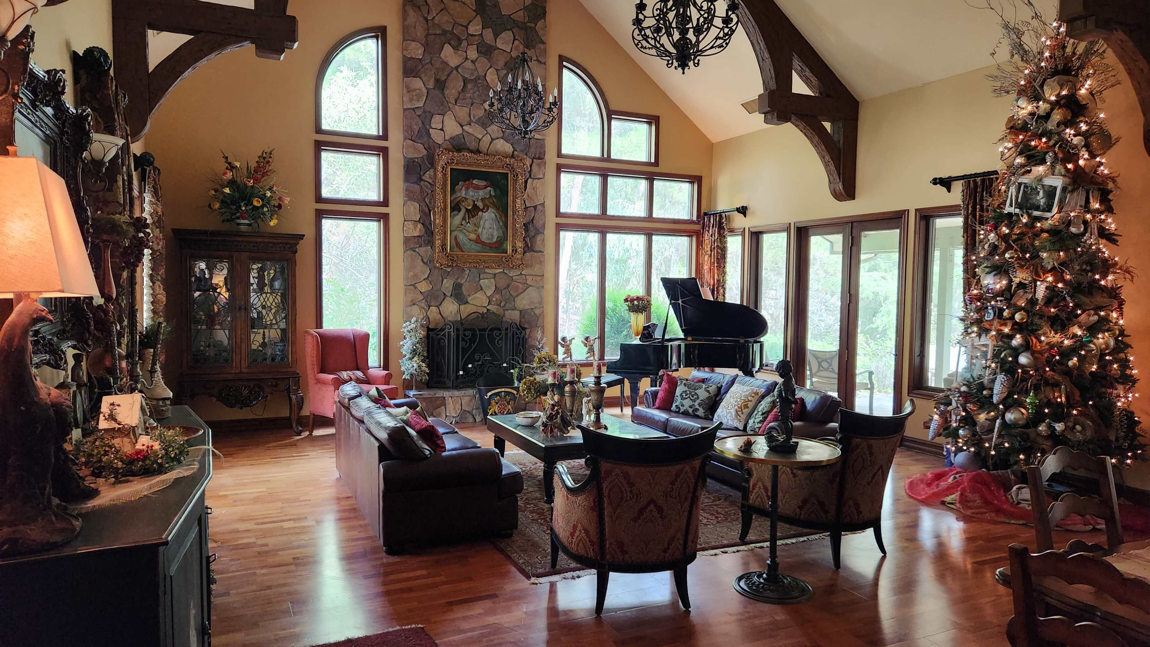 The image depicts a spacious living room with a tall stone fireplace, large windows, a grand piano, and a decorated Christmas tree in the corner.