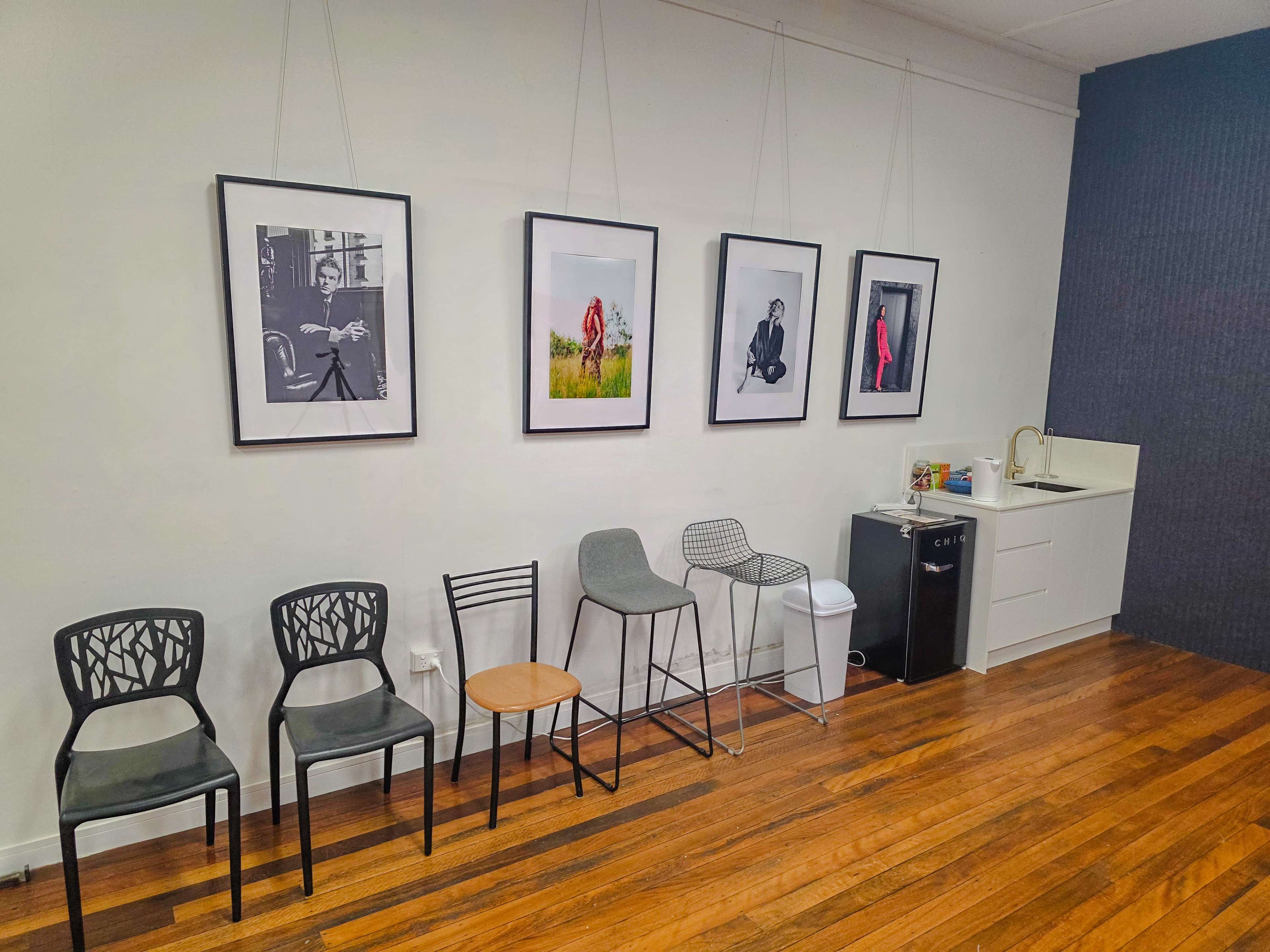 The scene features a wall with five framed black-and-white photographs, alongside a variety of chairs and a kitchenette area in a well-lit room with wooden flooring.