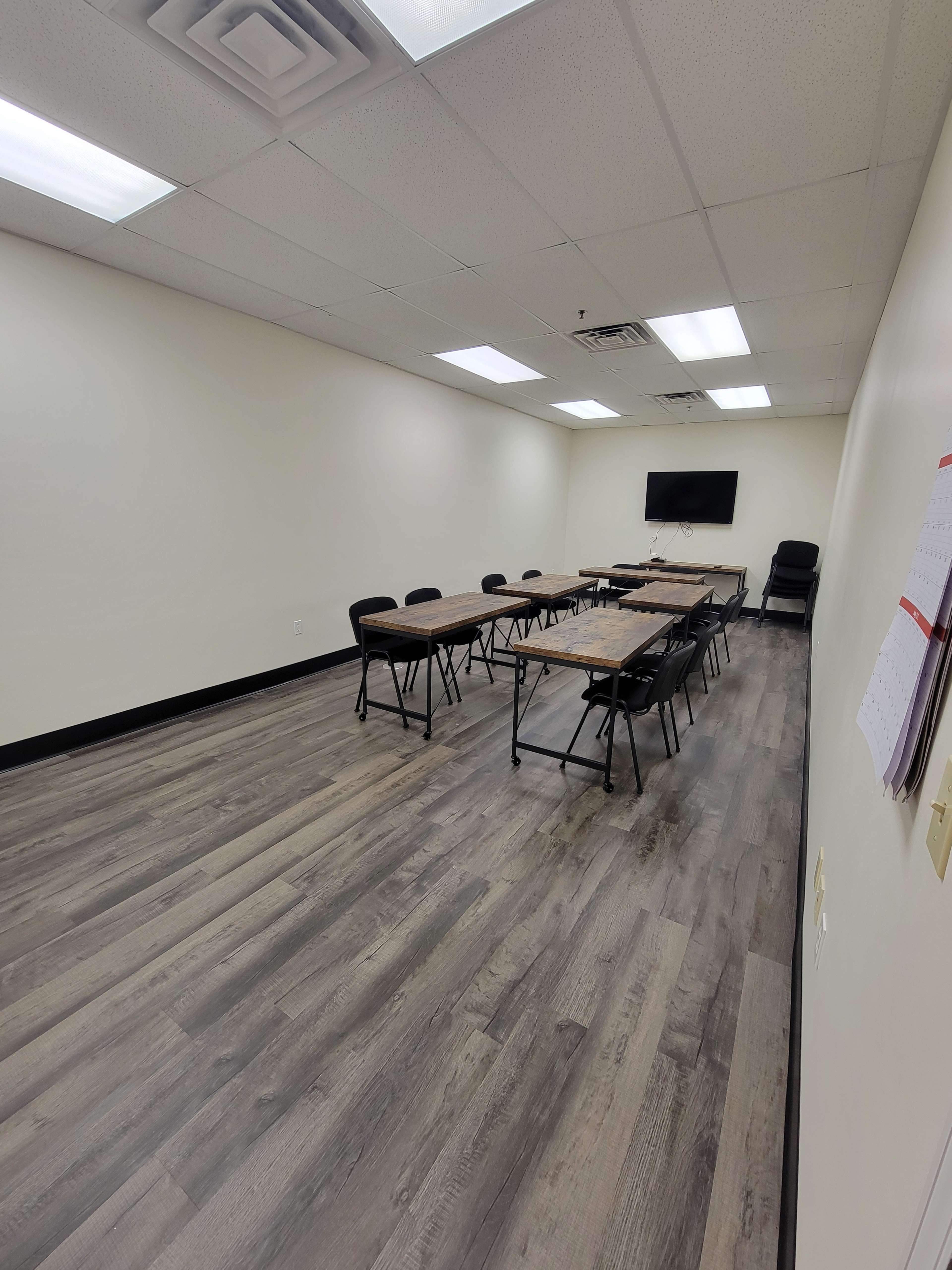 A sparsely furnished meeting room with a rectangular table, black chairs, and a wall-mounted screen.