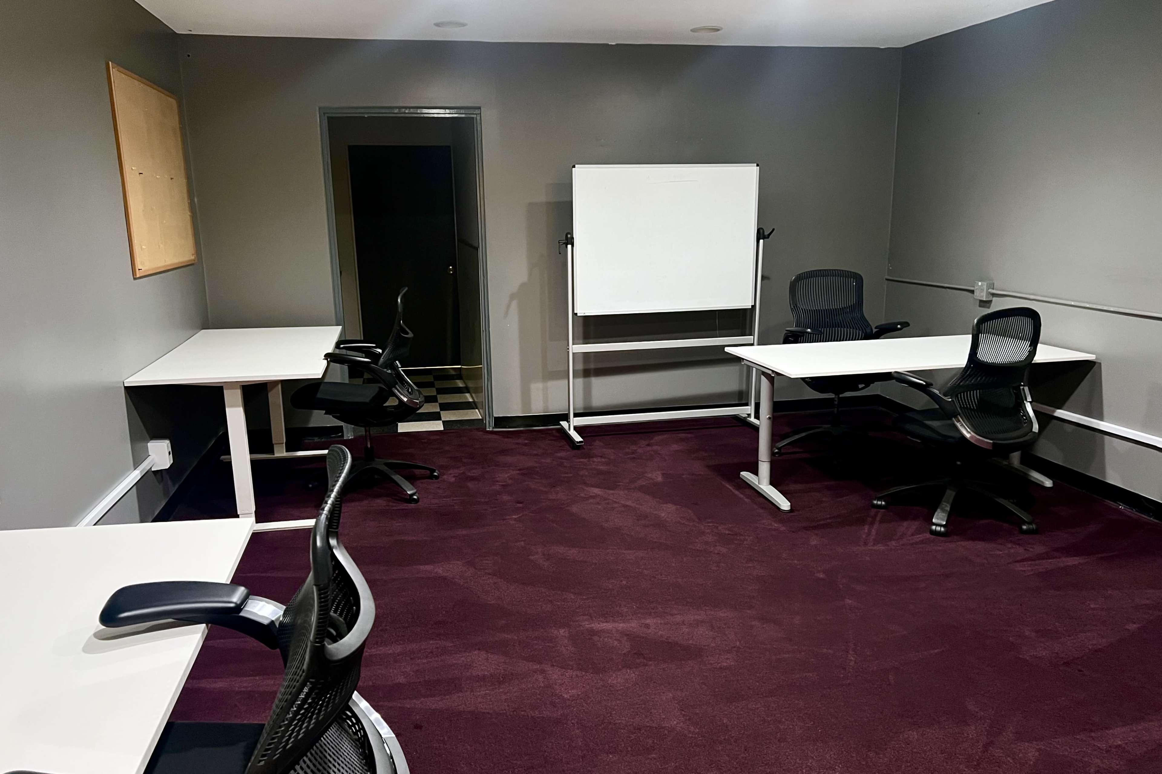 A small meeting room with four chairs and two white tables, featuring a whiteboard and a dark-colored carpeted floor.