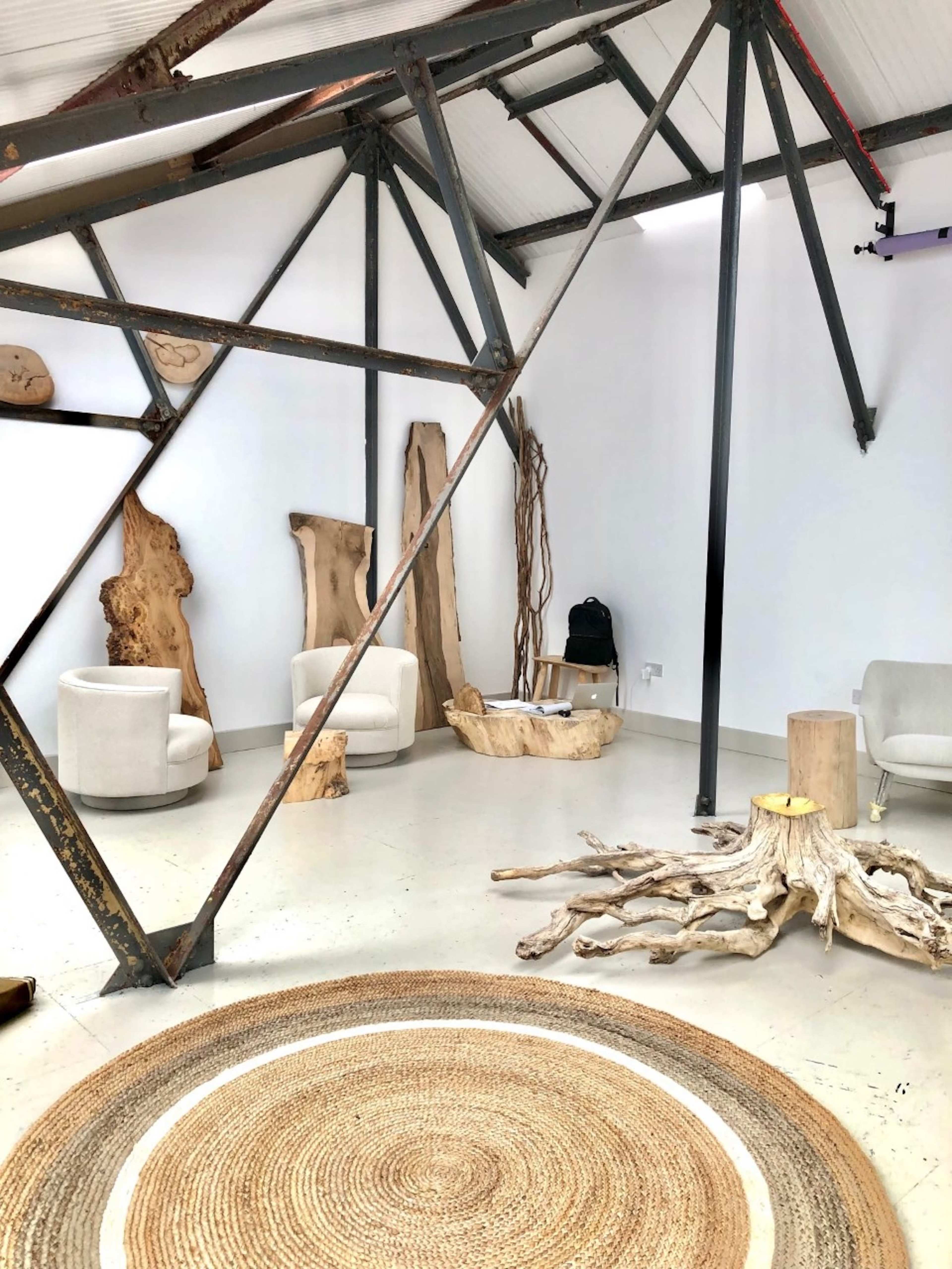 A modern, minimalist interior space featuring two white chairs, natural wood decor pieces, and a large circular rug on a concrete floor, all framed by visible metal beams.