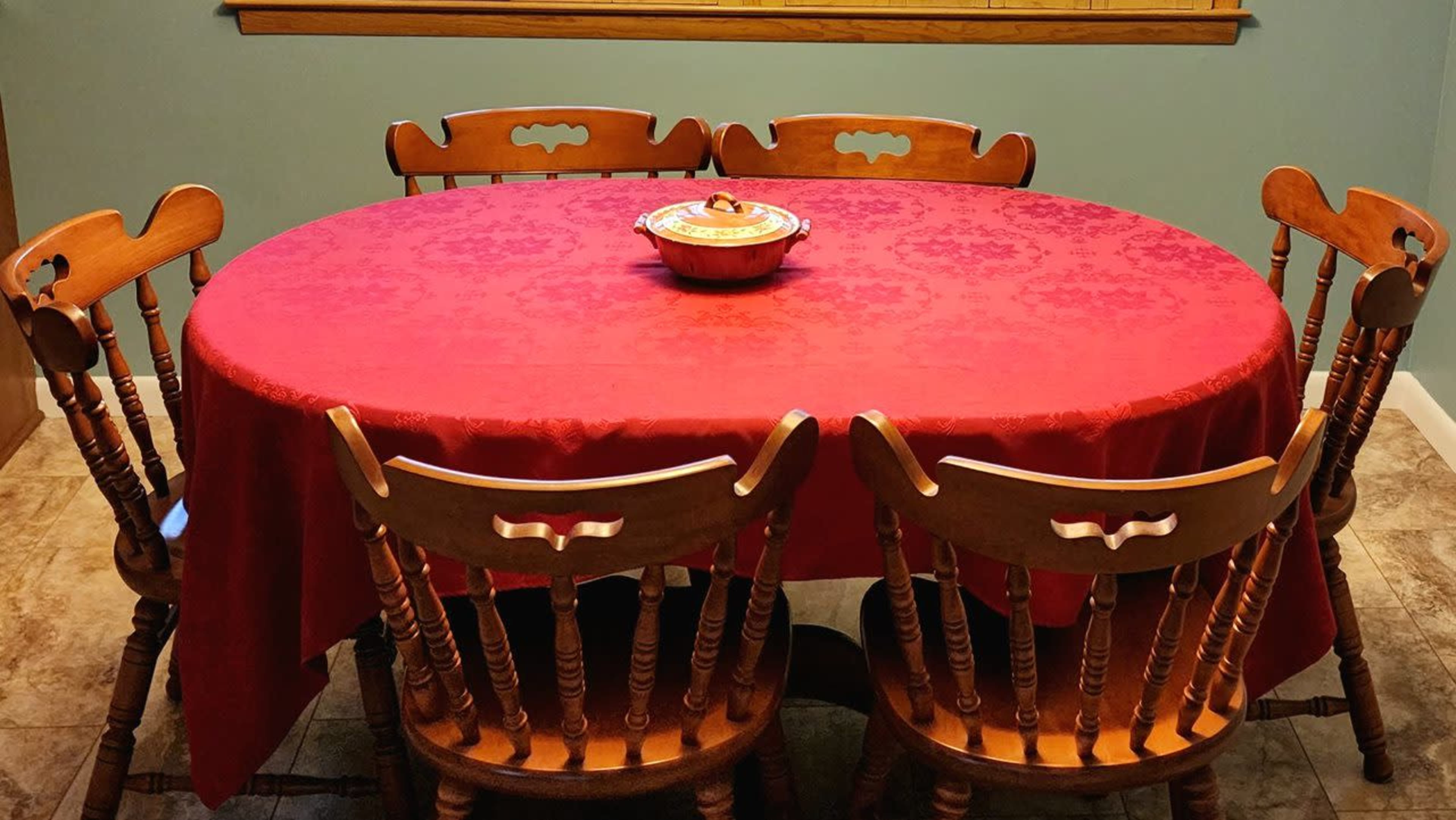 An oval dining table with a red tablecloth is surrounded by six wooden chairs, with a dish sitting in the center.