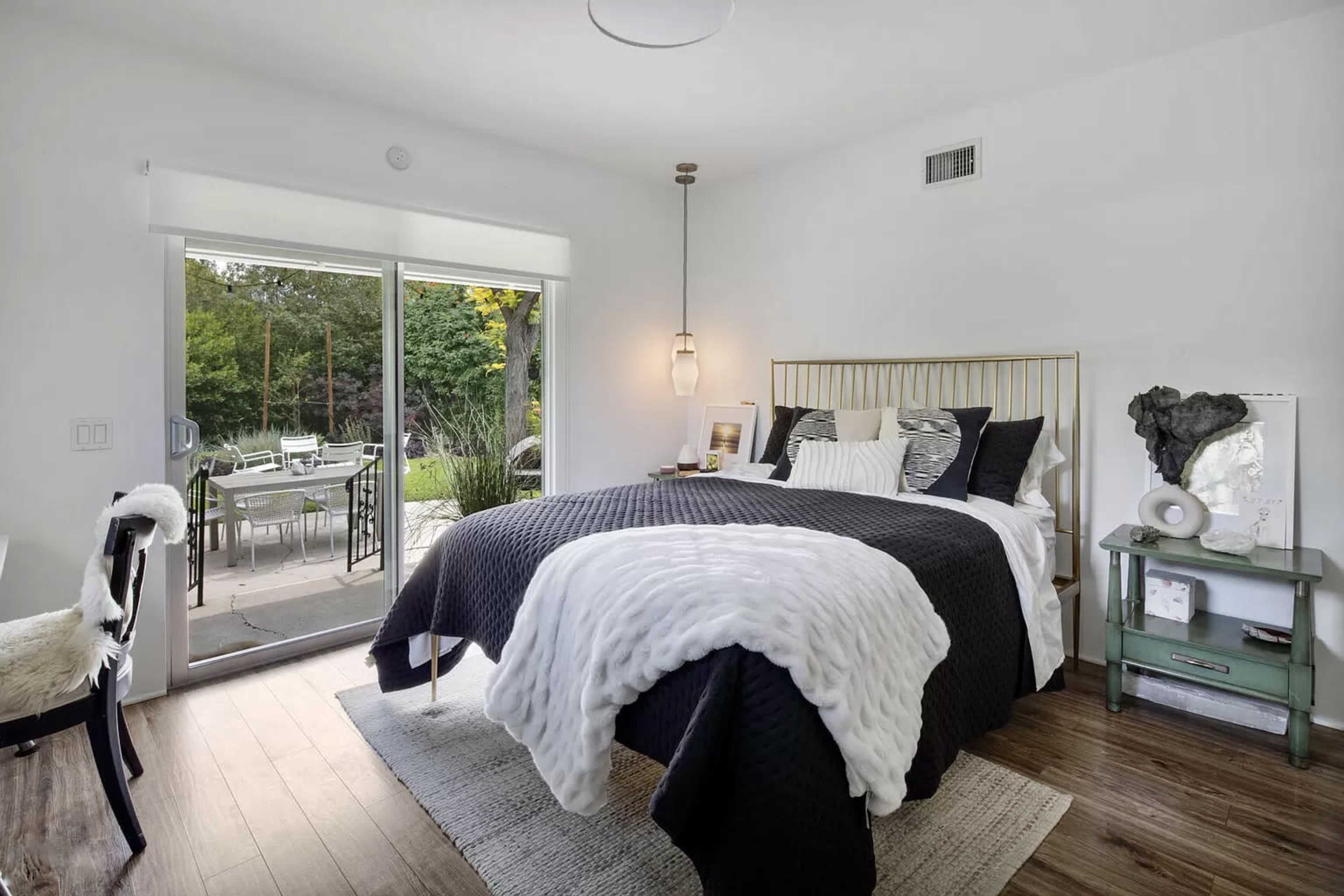 A modern bedroom features a large bed with black and white bedding, situated beside sliding glass doors that open to an outdoor patio with furniture.