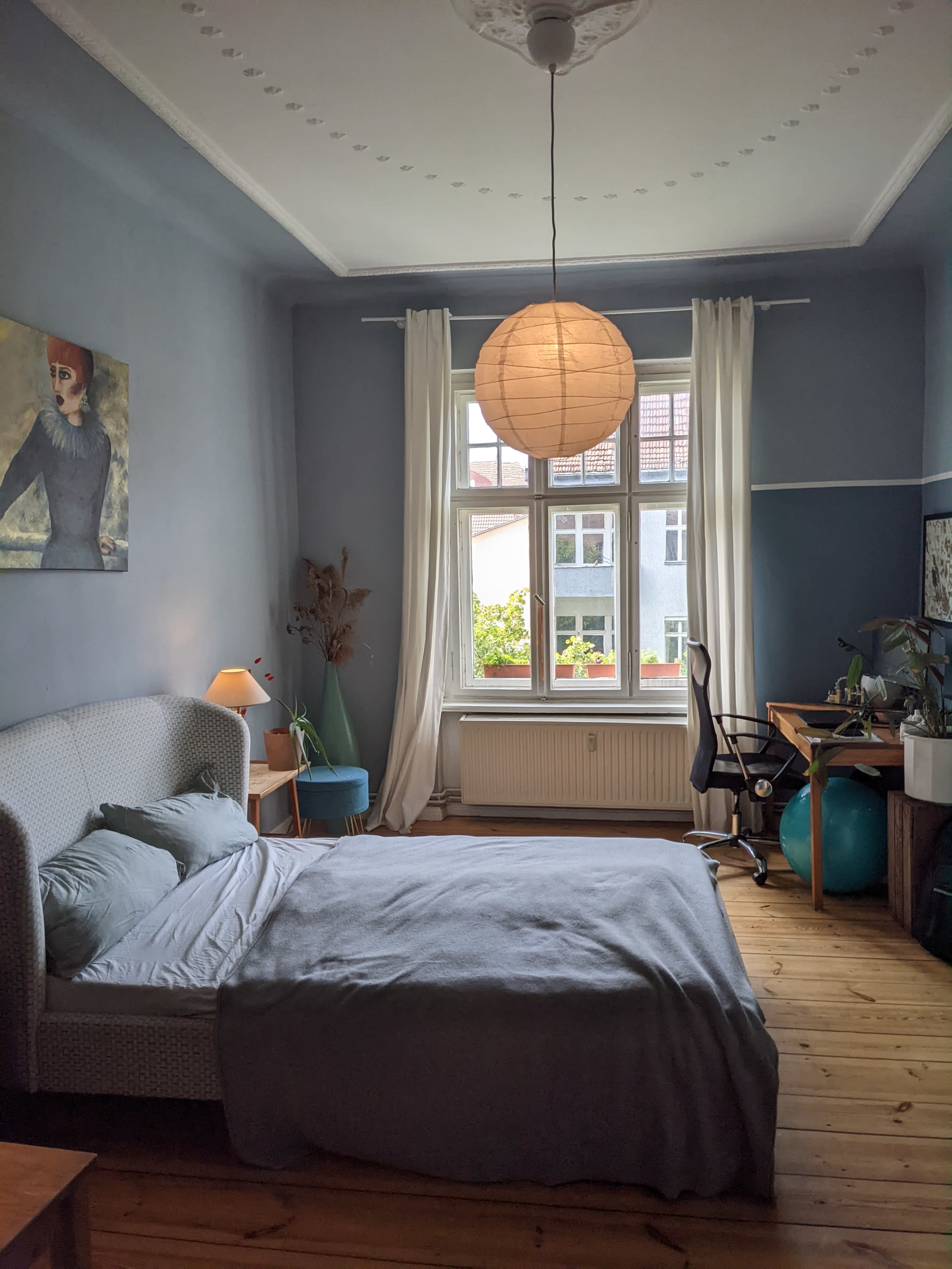 The image shows a cozy bedroom with a bed, a desk with a chair, and a large window allowing natural light to enter.