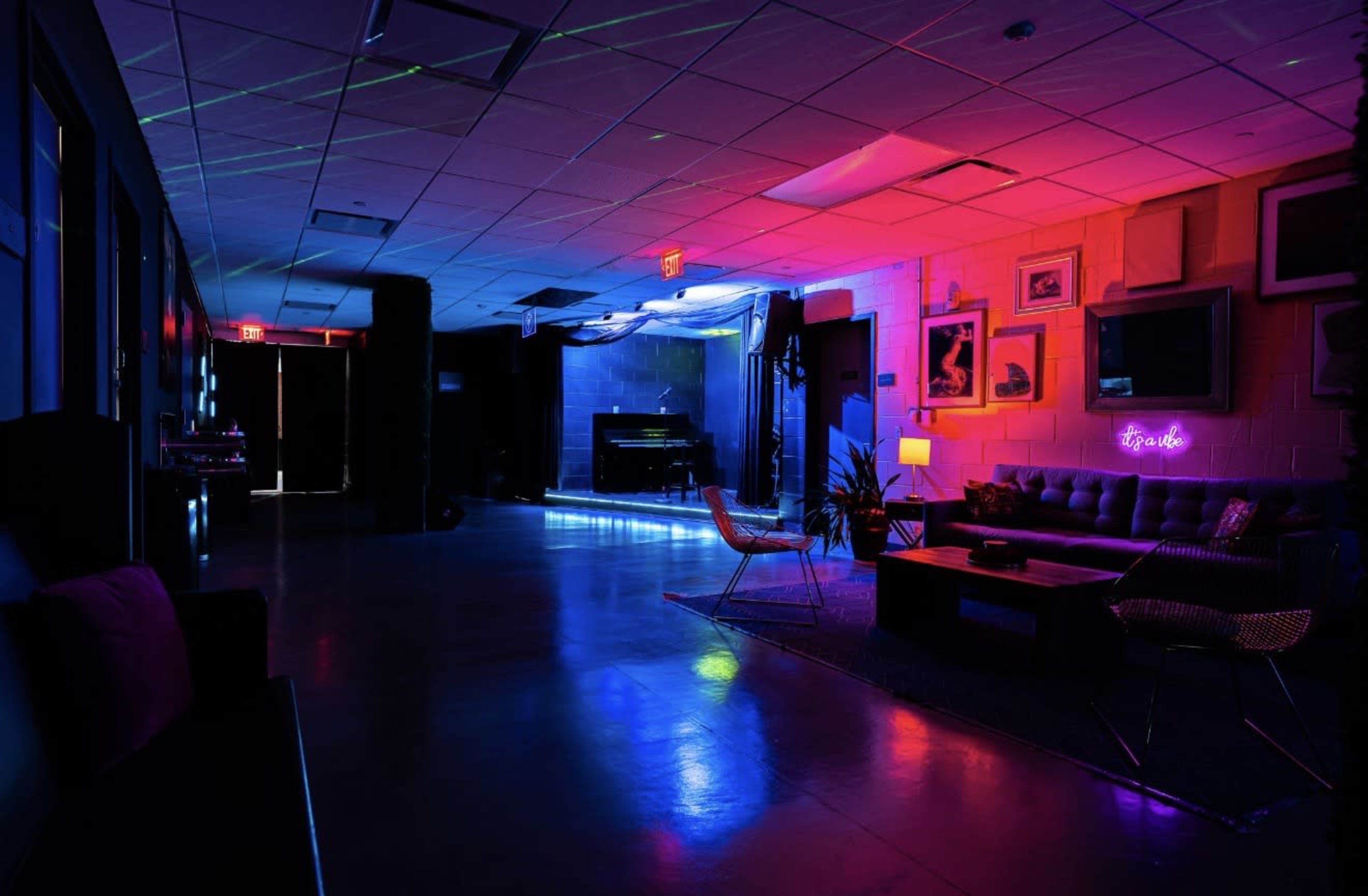 The image shows a dimly lit lounge area featuring colorful neon lights, a piano, and various seating arrangements.