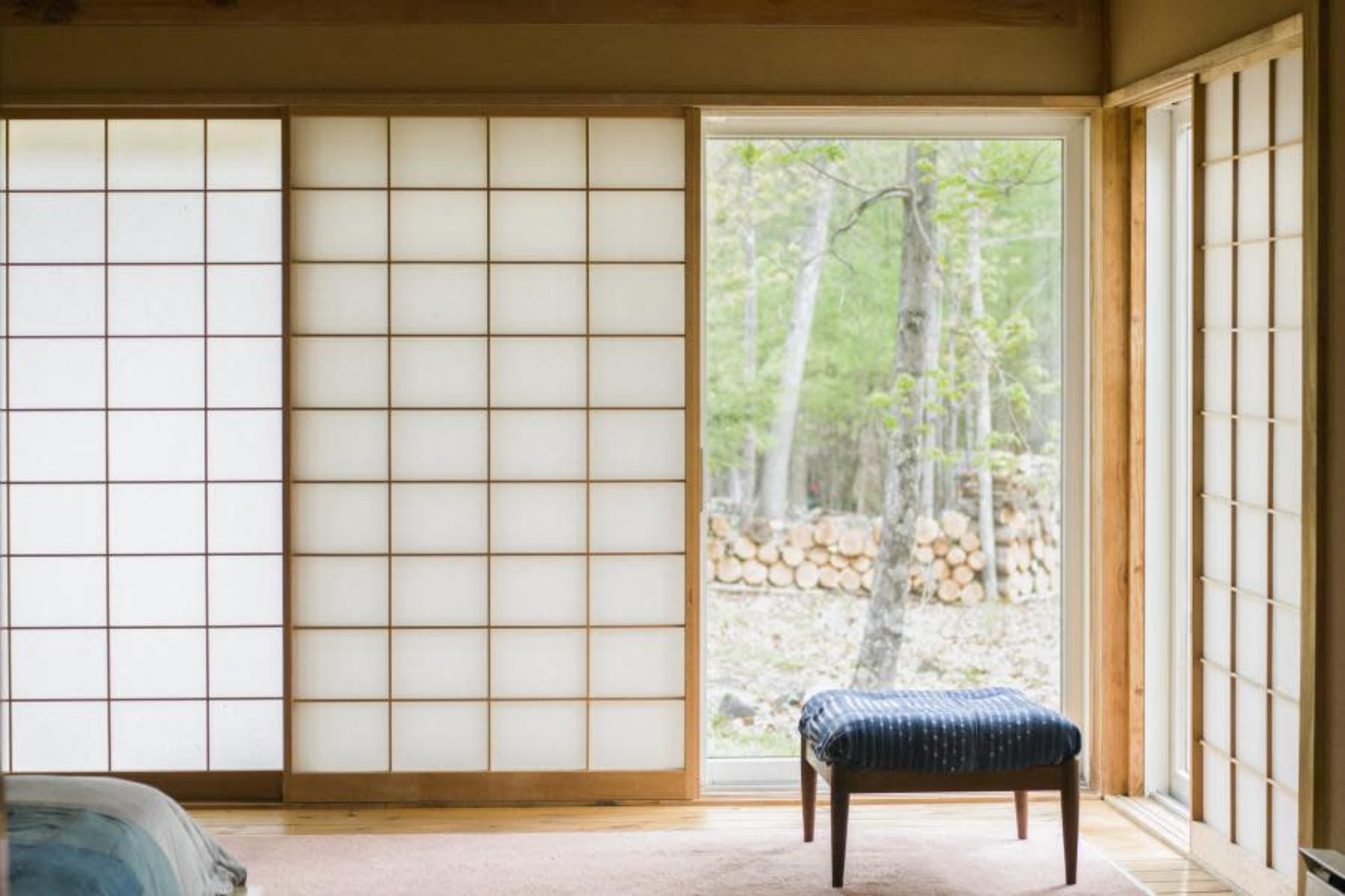 A minimalist interior features sliding shoji screens, a small wooden stool, and a view of a forested area through an open door.
