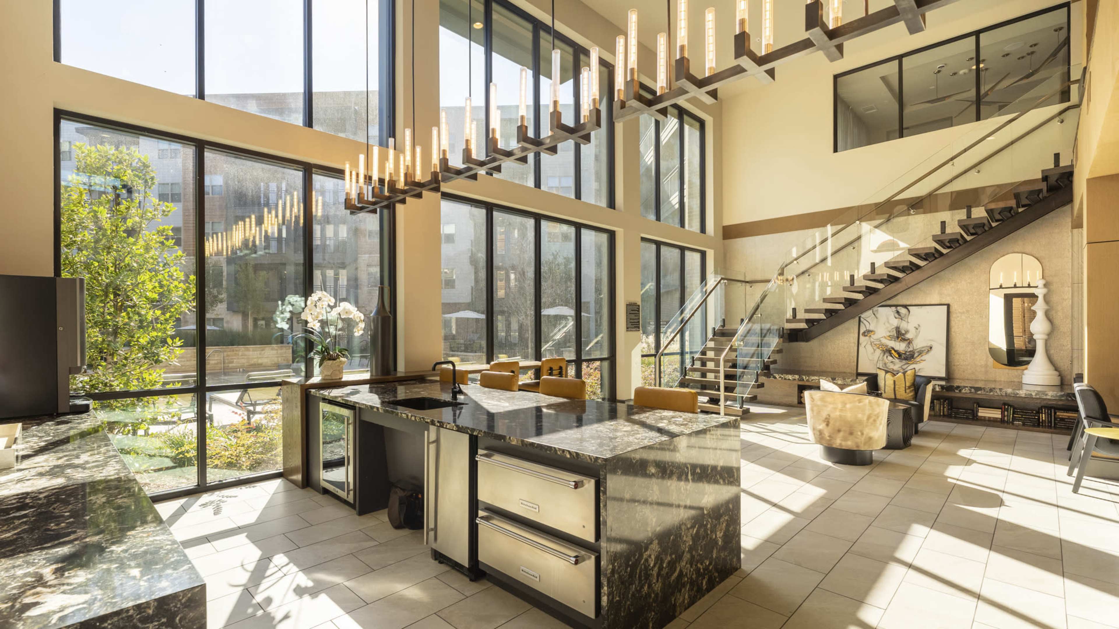 A modern open-concept living space featuring large windows, a marble kitchen island, and a staircase leading to an upper level.