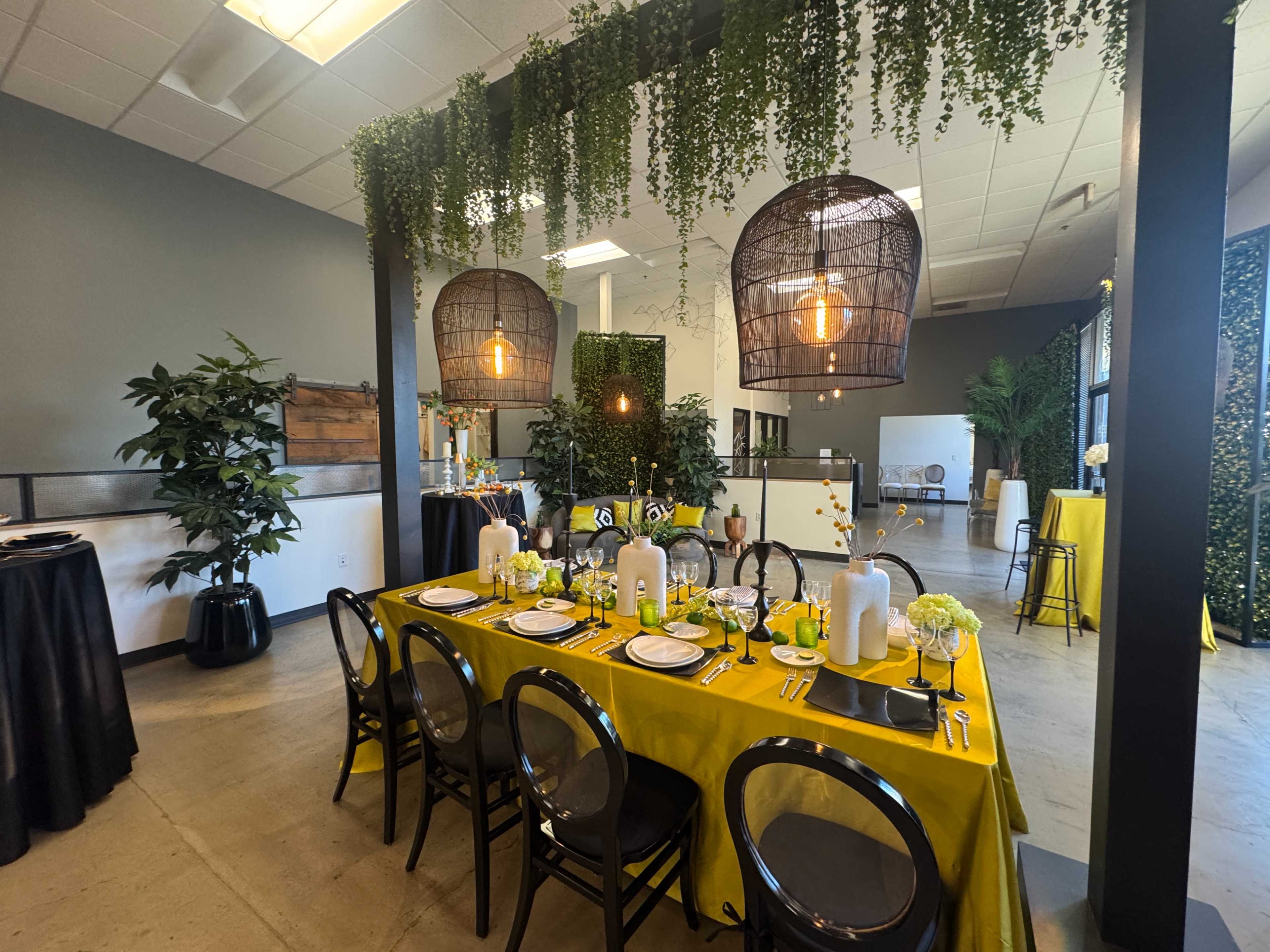The image shows a decorated dining space with a long table set with yellow linens, surrounded by black chairs, and adorned with plants and hanging light fixtures.