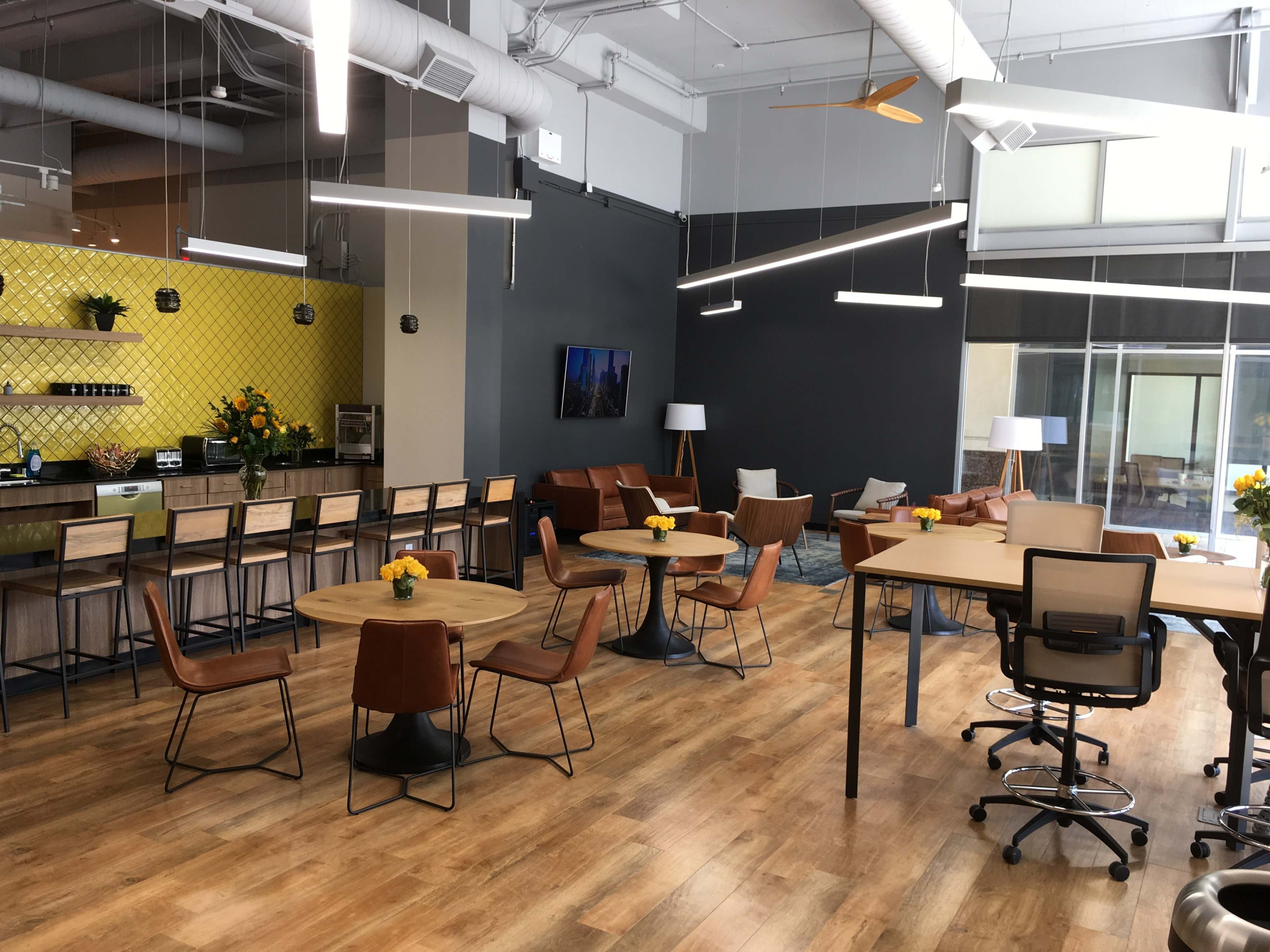 A modern office space with wooden flooring, featuring tables and chairs, a coffee bar, and a small seating area with plants and decorative lighting.