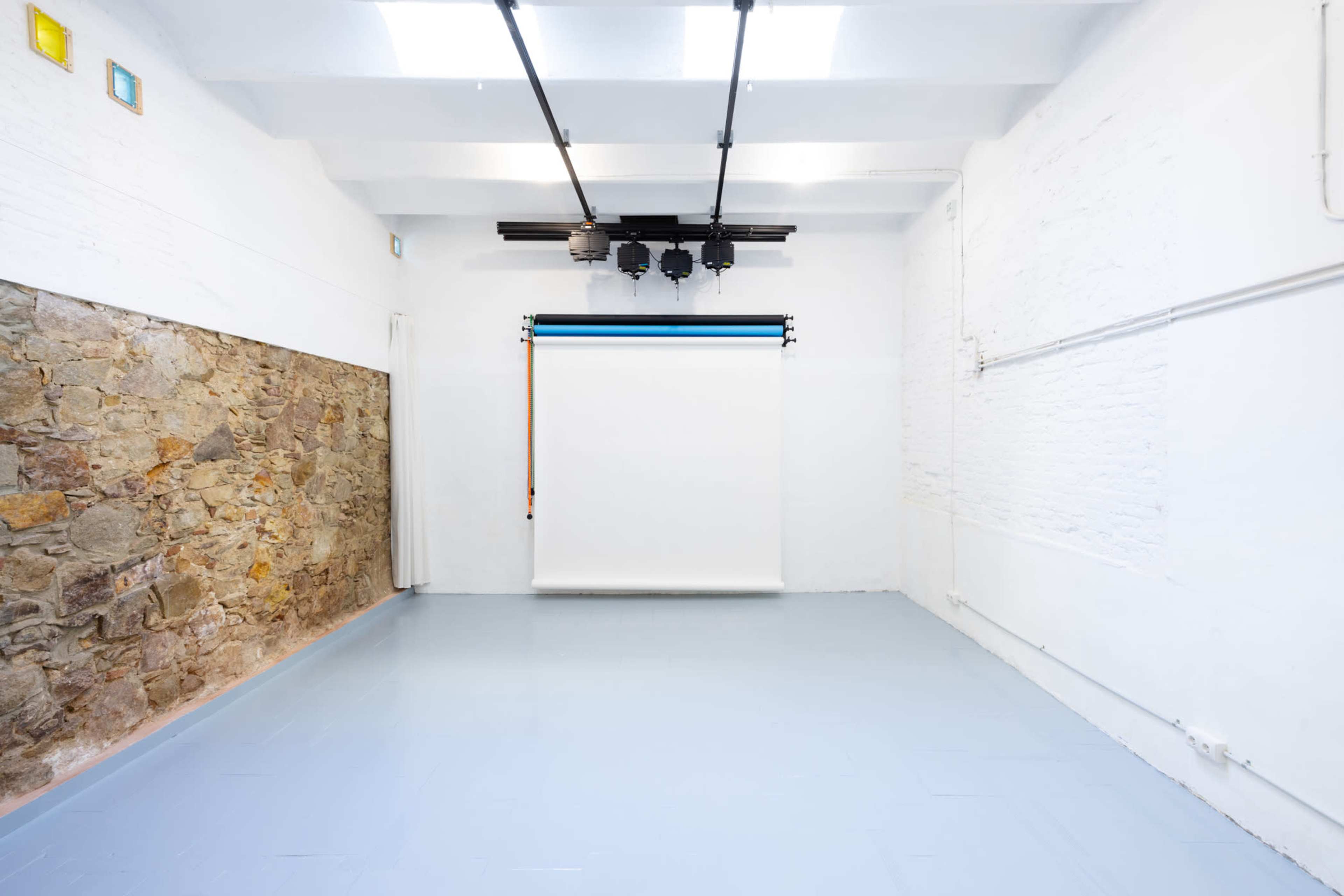 The image shows a minimalist studio space with a white wall, a section of exposed stone, and a retractable white backdrop.