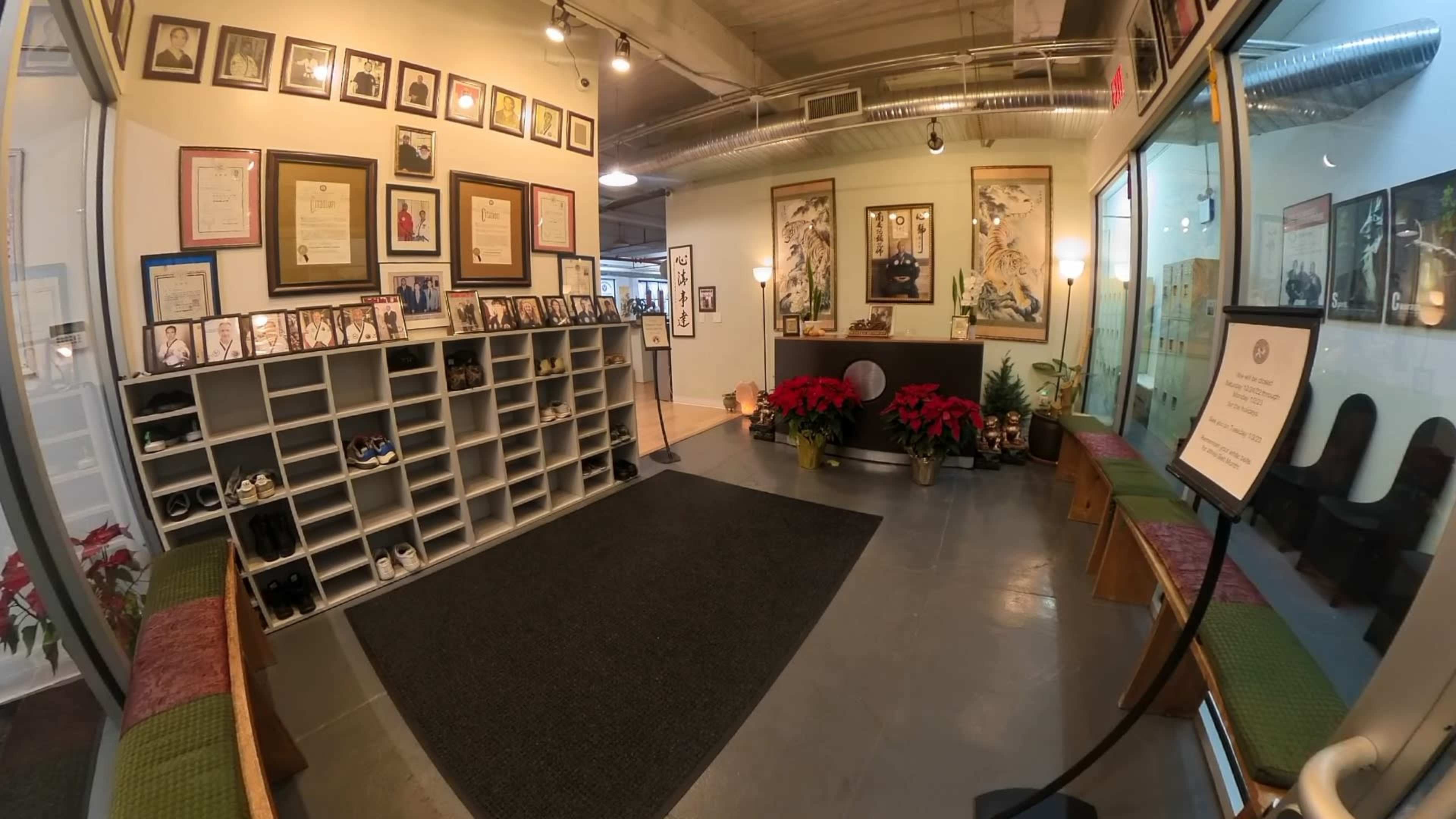 The image shows a brightly lit reception area with several framed documents and photographs on the walls, a shoe cubby, potted plants, and a seating area.