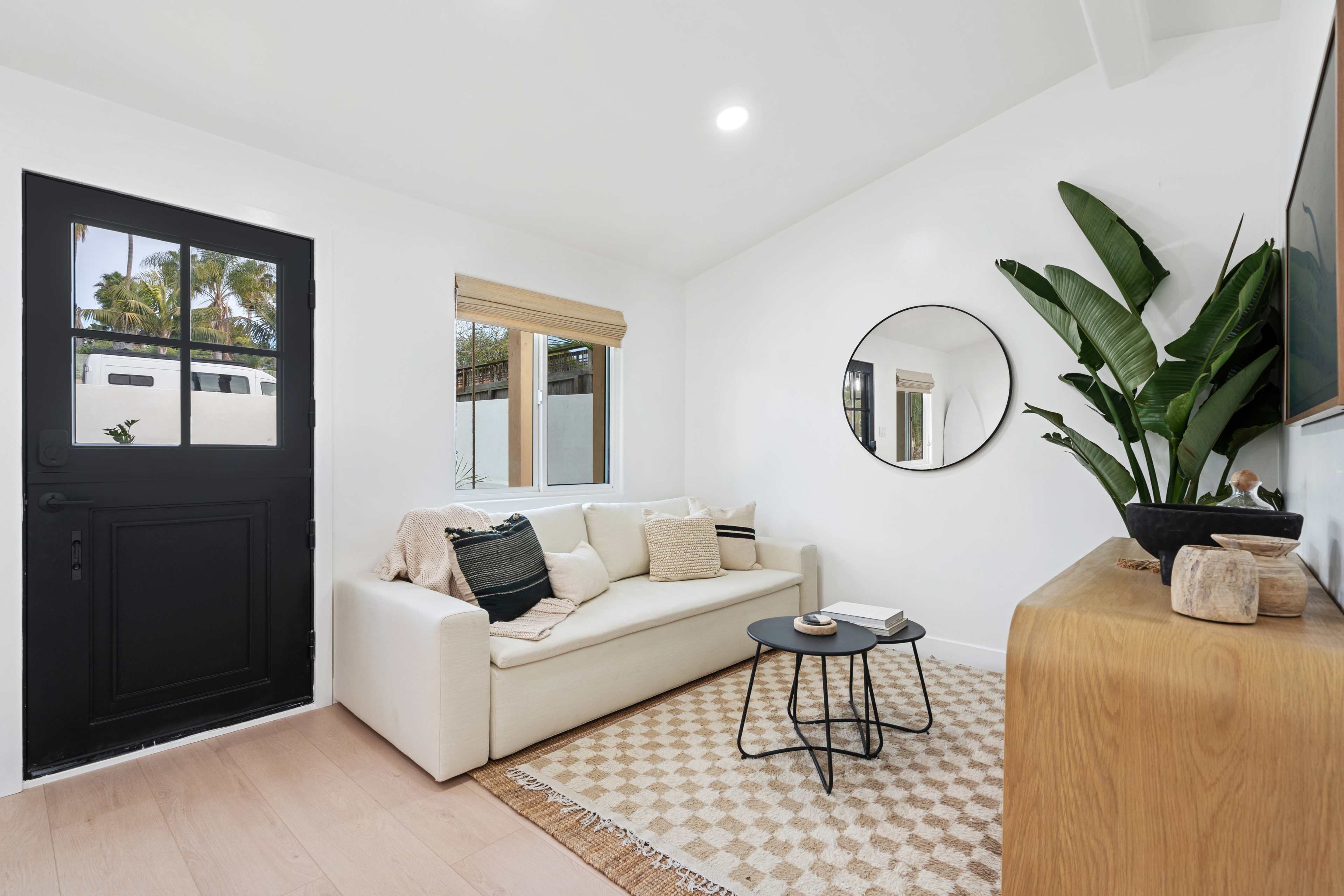 A bright living room with a white sofa, a round mirror, a black side table, and a plant next to a wooden cabinet.