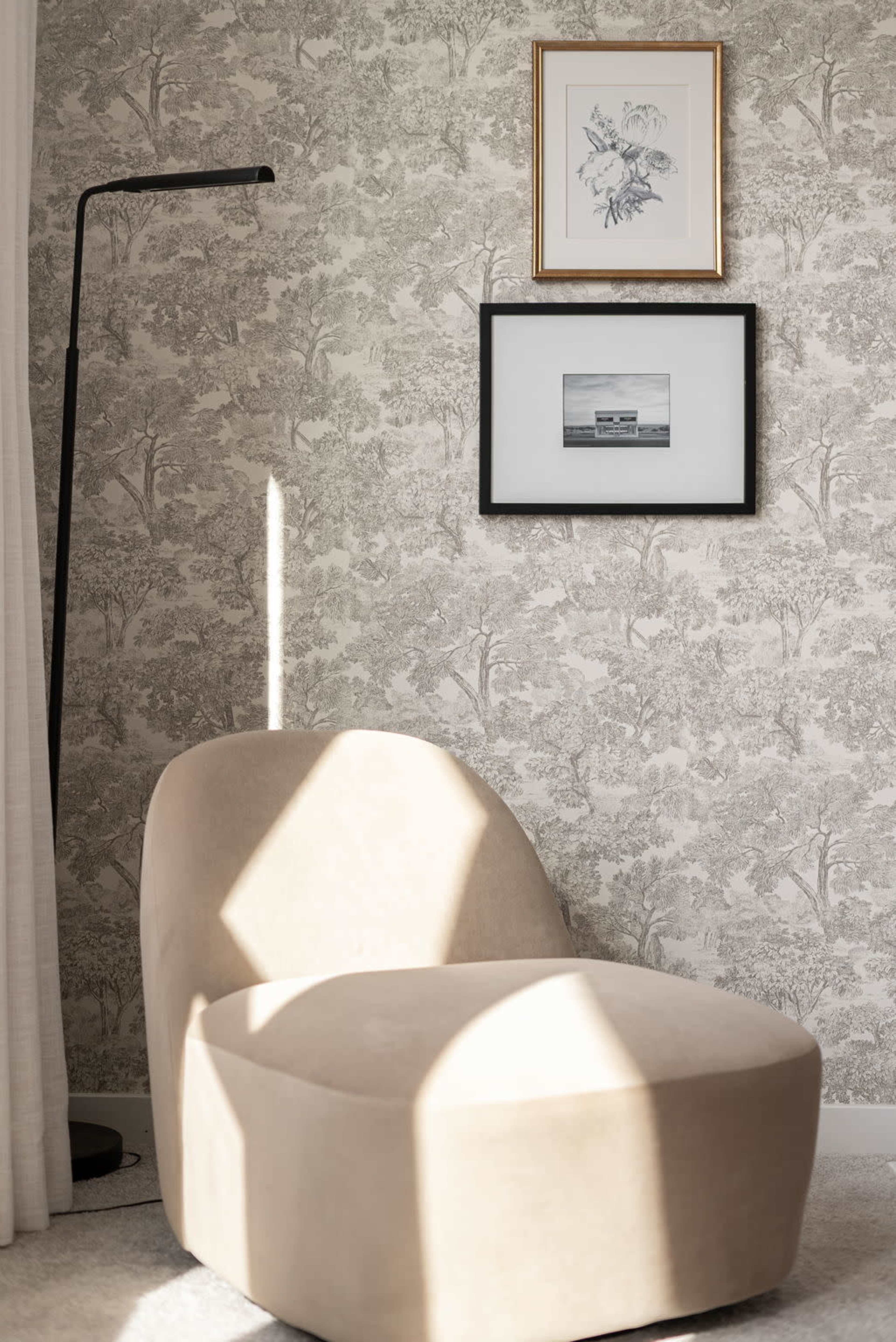 A curved, beige lounge chair sits next to a floor lamp on a patterned wallpaper background, adorned with two framed artworks above.