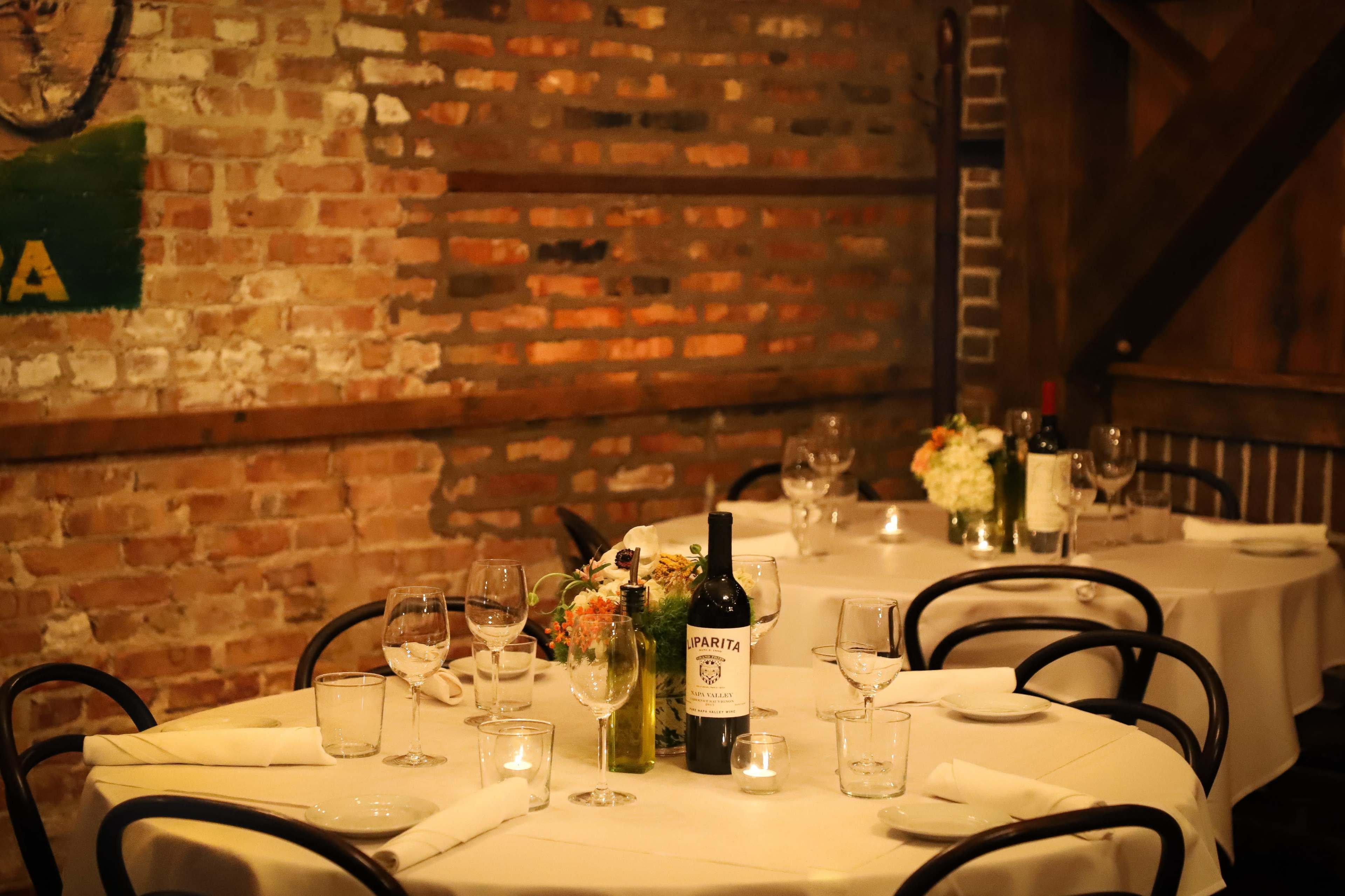 A cozy restaurant interior features a set table with white linens, wine bottles, and small flower arrangements against a brick wall backdrop.