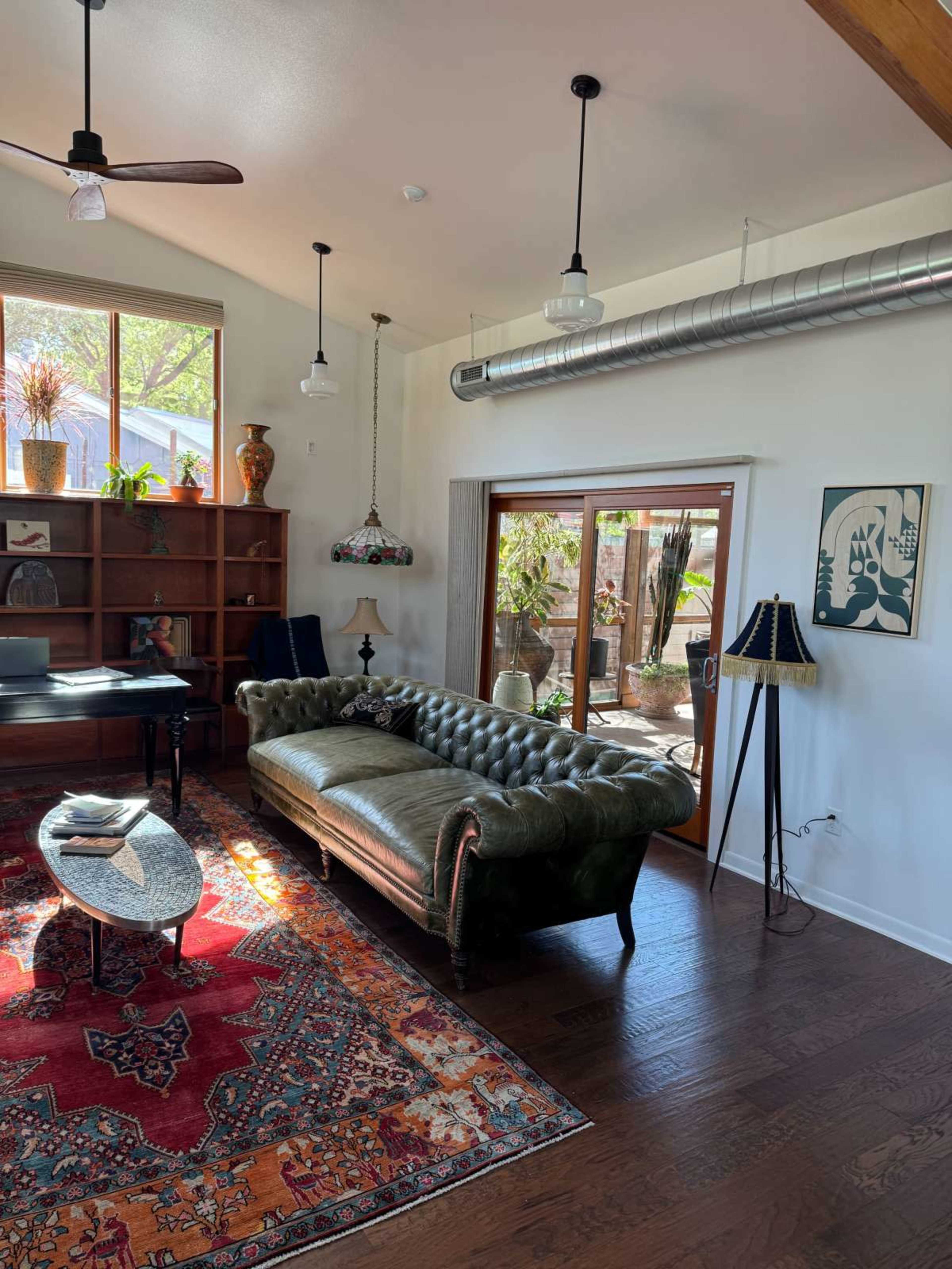 The image shows a cozy living room with a green leather chesterfield sofa, a wooden bookshelf, and large windows allowing natural light to illuminate the space.