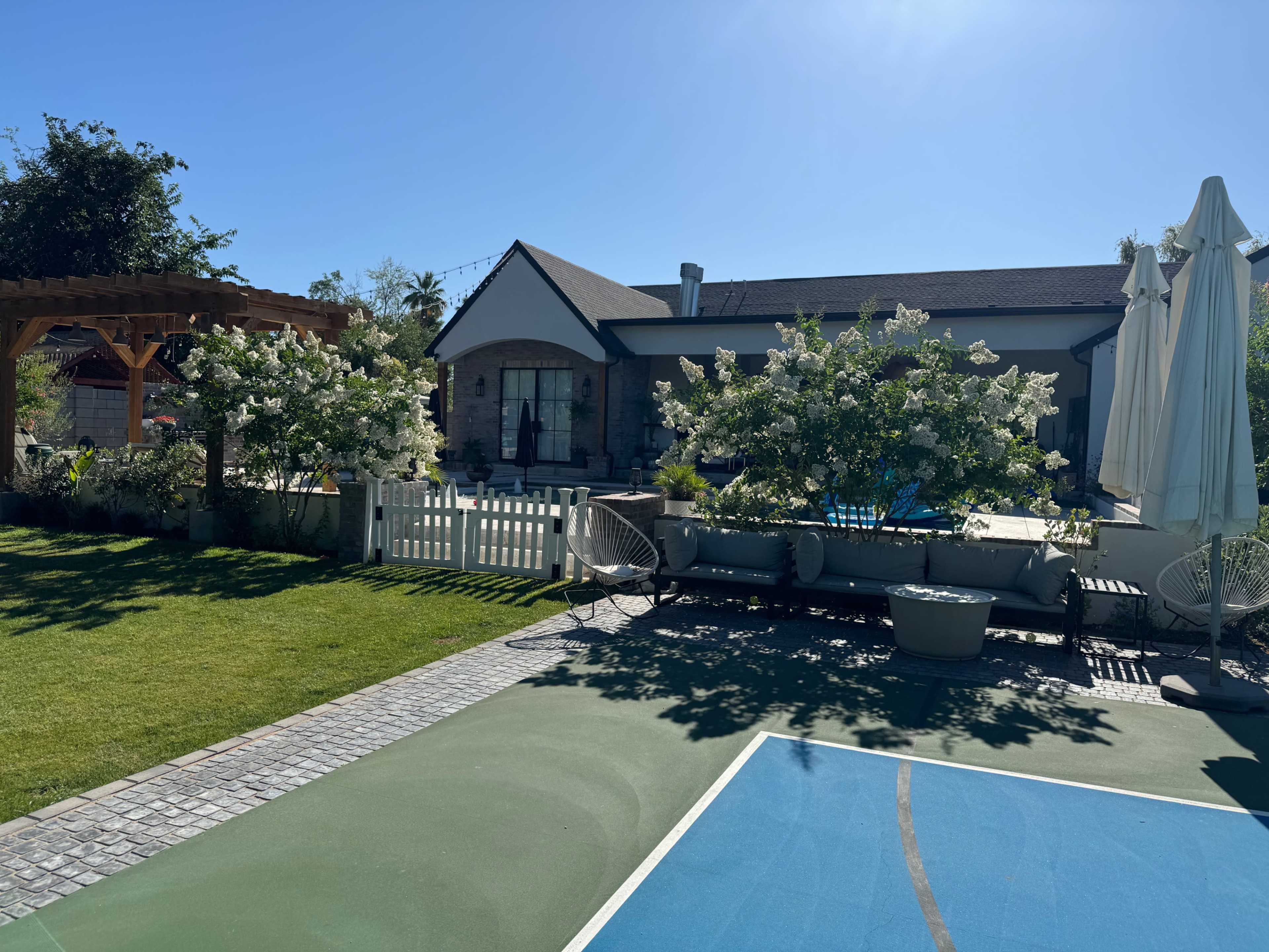 A backyard featuring a swimming pool, a basketball court, and flowering shrubs alongside a modern house.