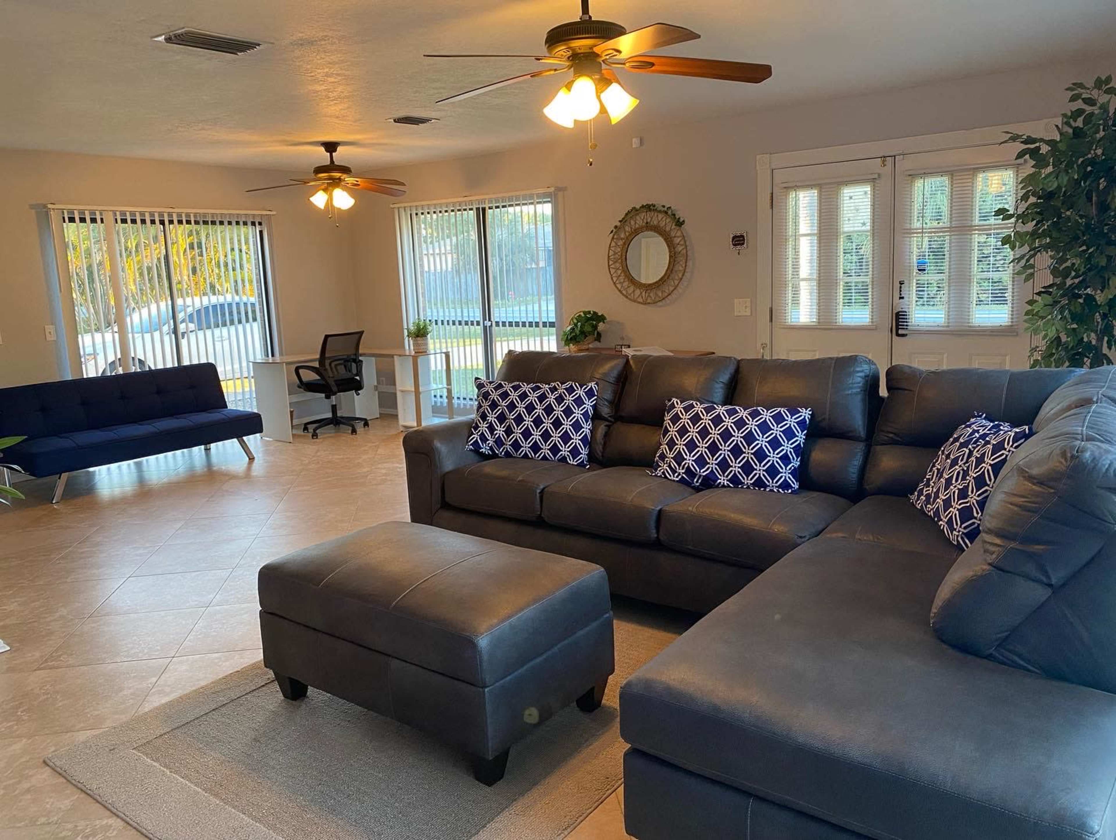 The room features a sectional sofa with blue and white pillows, a coffee table, and a desk area, along with two ceiling fans and large windows that provide natural light.