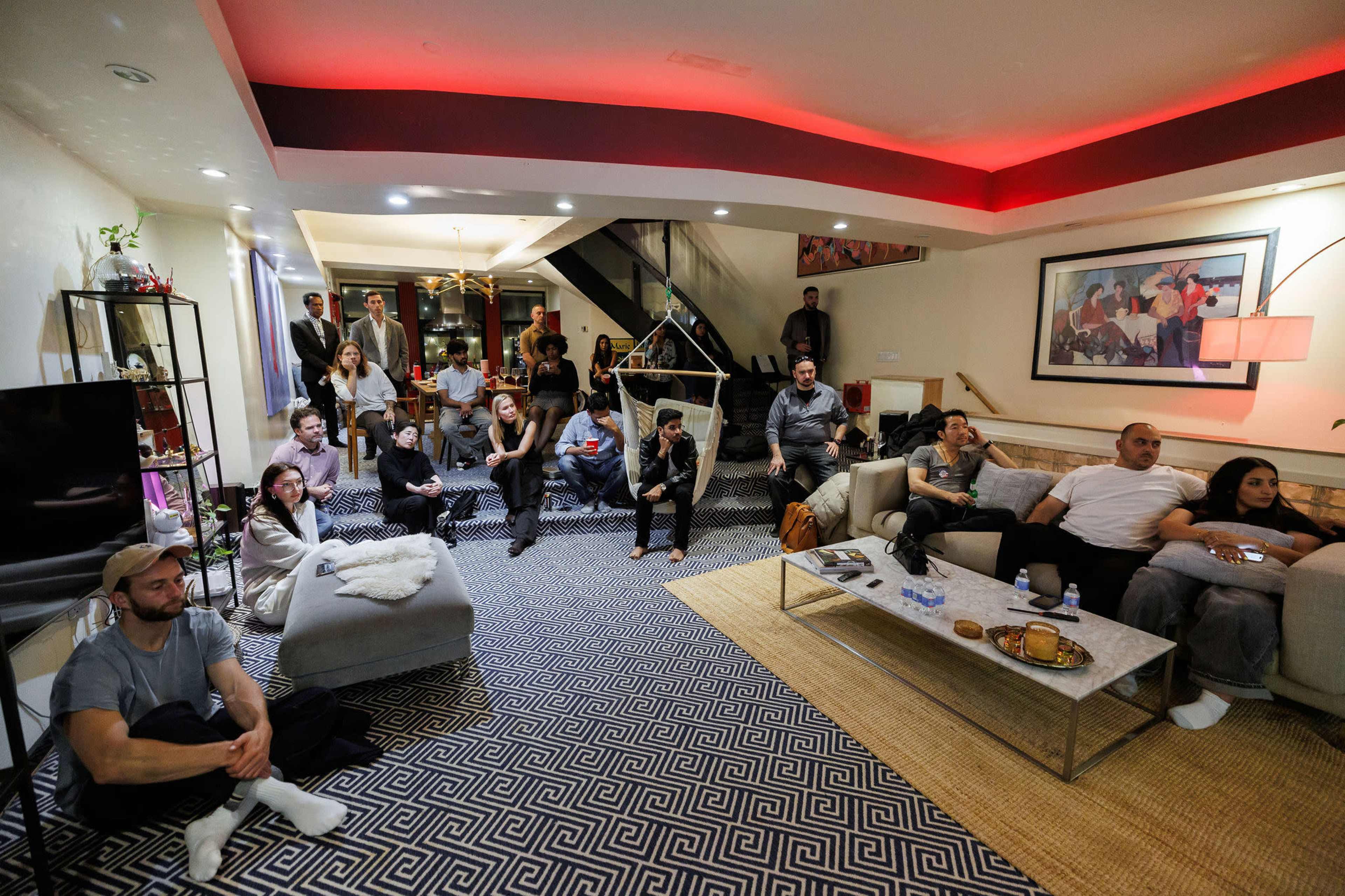 A group of people sits in a living room, watching something intently on a large screen.
