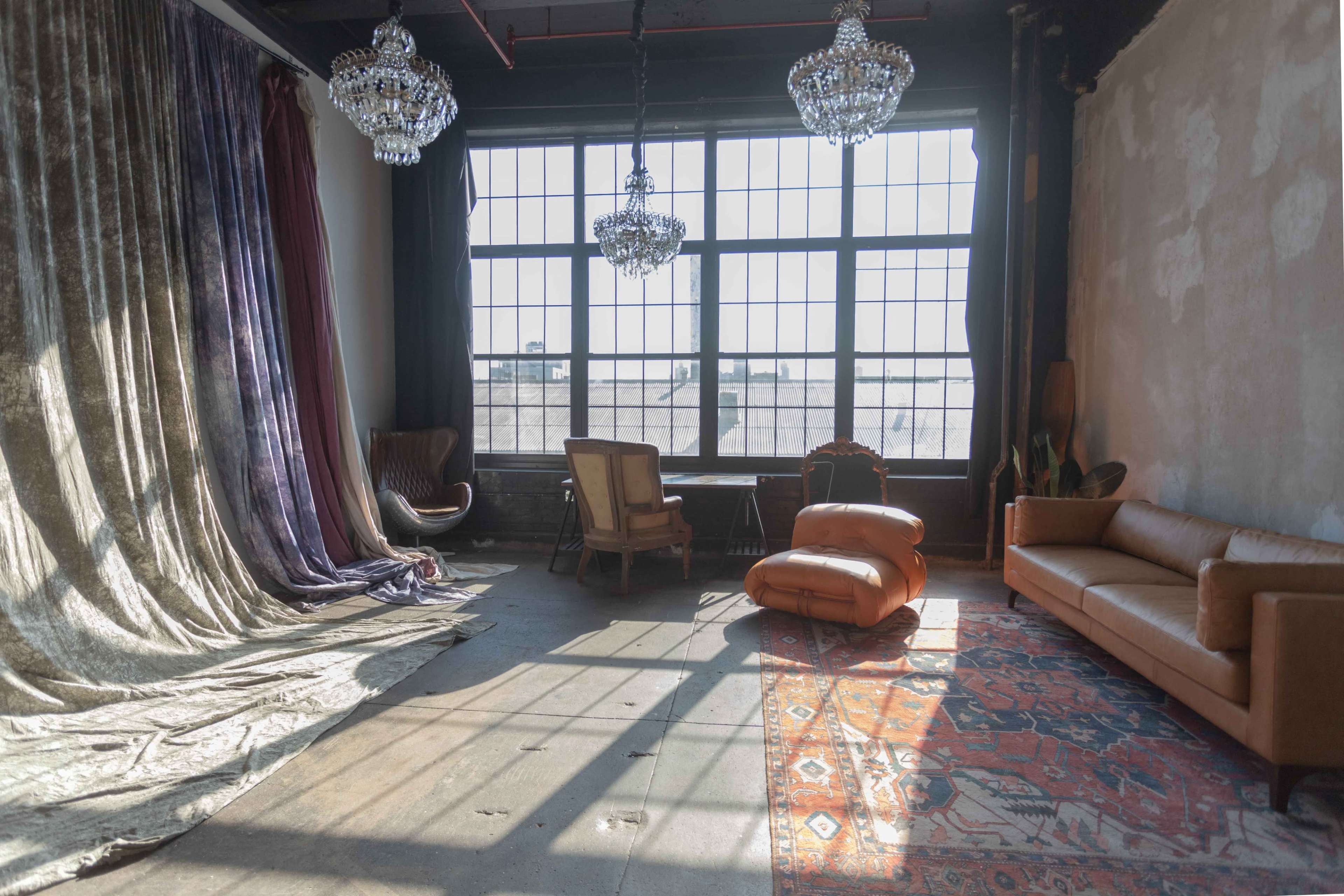 A spacious room features large windows, two chandeliers, a Persian rug, a leather sofa, and various seating arrangements against textured walls.