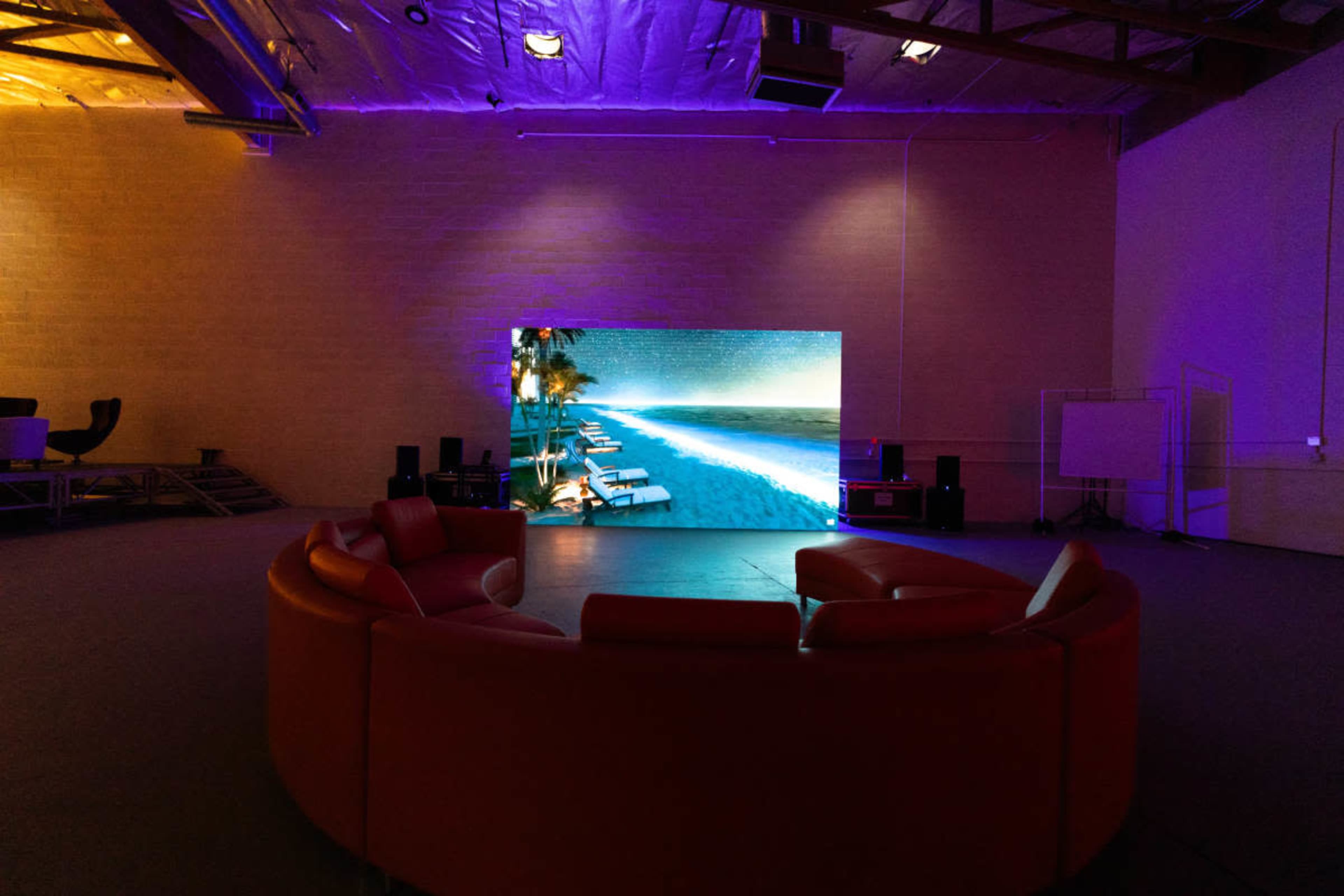 A circular red sofa faces a large screen displaying a beach scene in a dimly lit room.