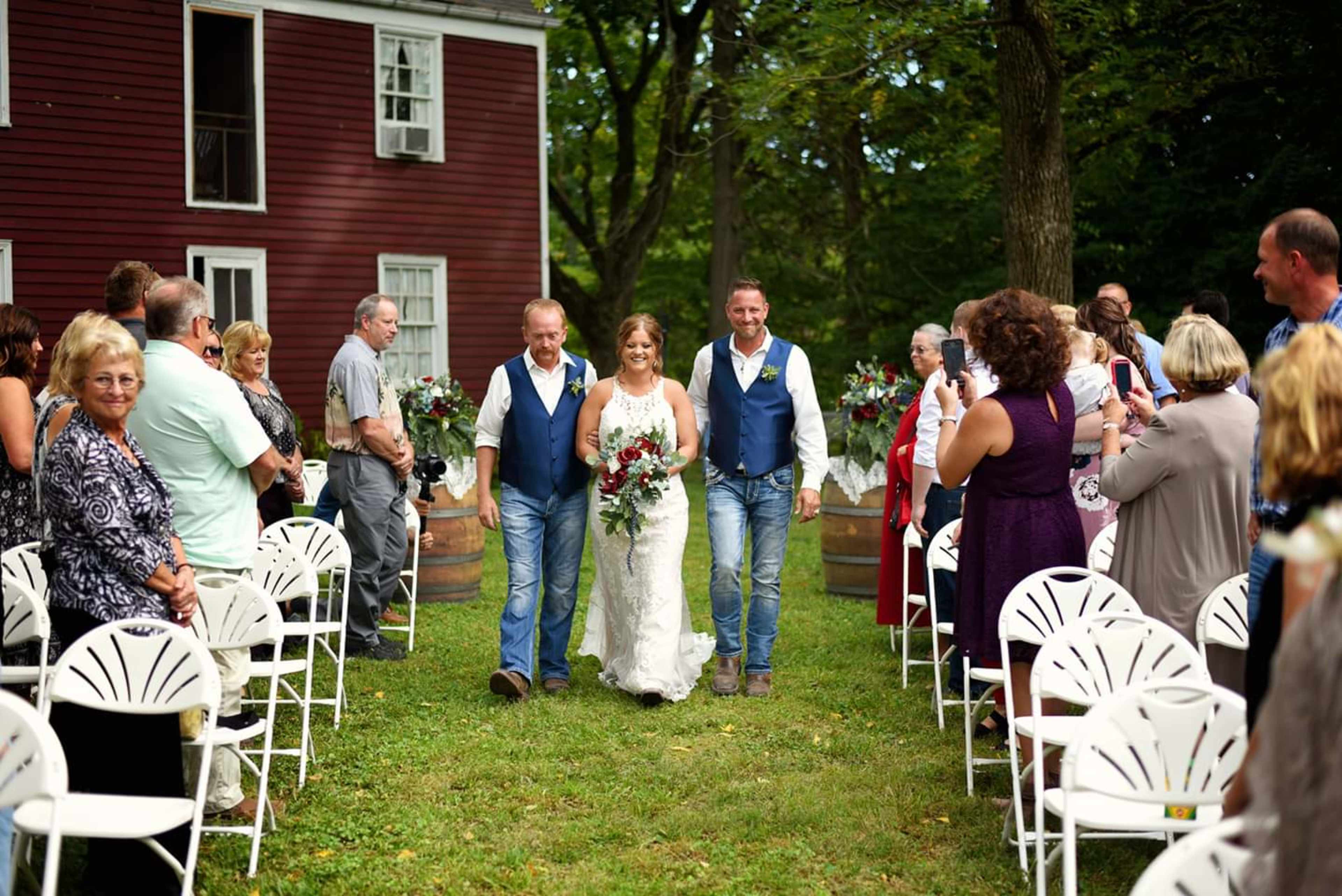 A bride walks down an outdoor aisle flanked by two men, flanked by guests seated on white chairs arranged in a grassy area.