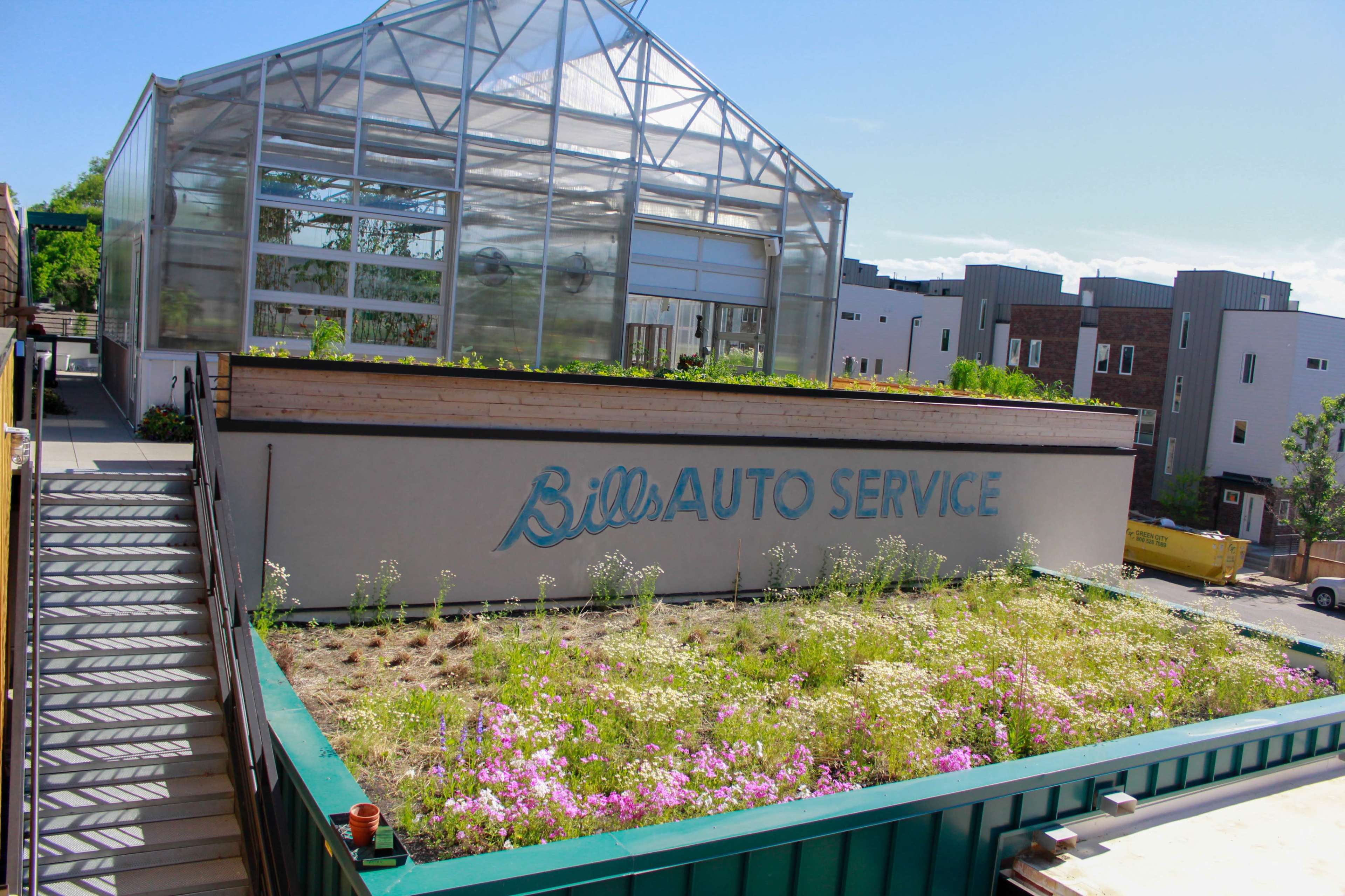 A green roof with flowering plants is situated above the sign for Bill's Auto Service, adjacent to a glass greenhouse and modern buildings.