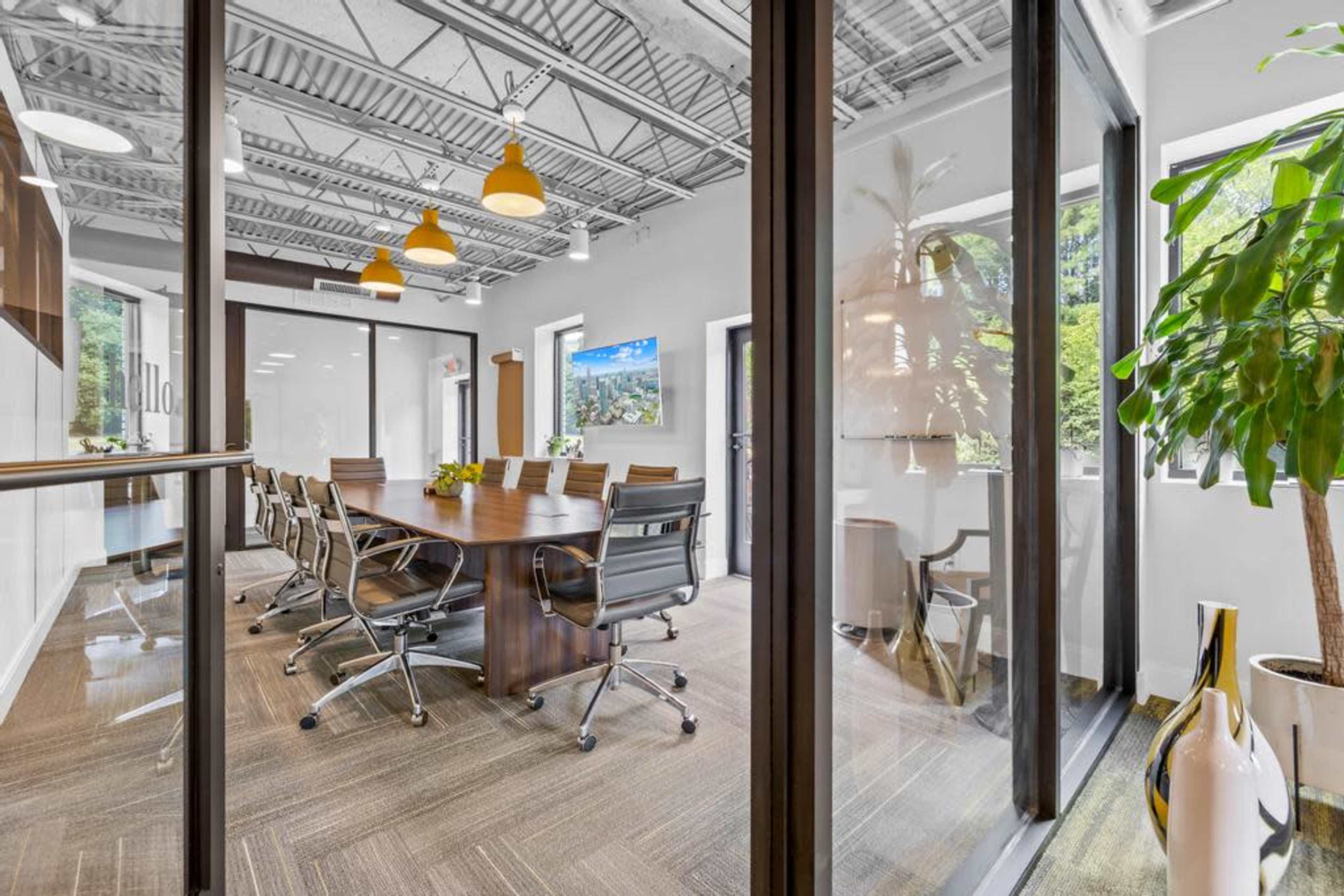 A modern conference room features a large table surrounded by chairs, with glass walls, a television, and plants in the background.