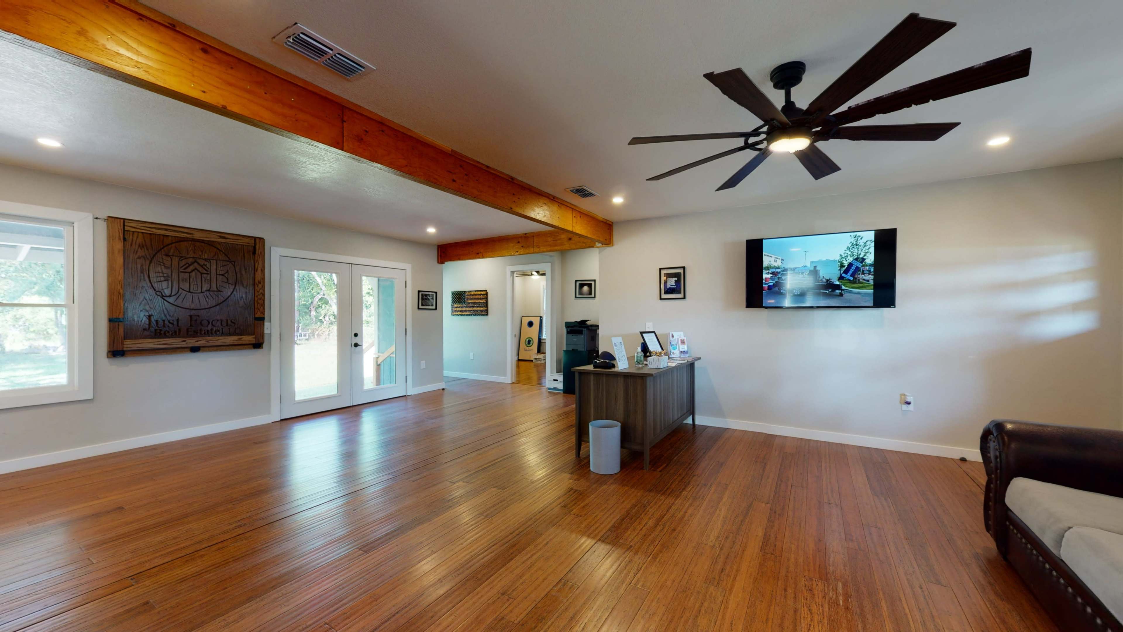 The image shows a spacious interior room with wooden beams, a ceiling fan, a television mounted on the wall, and light-colored walls, featuring a sofa and a table with various items.
