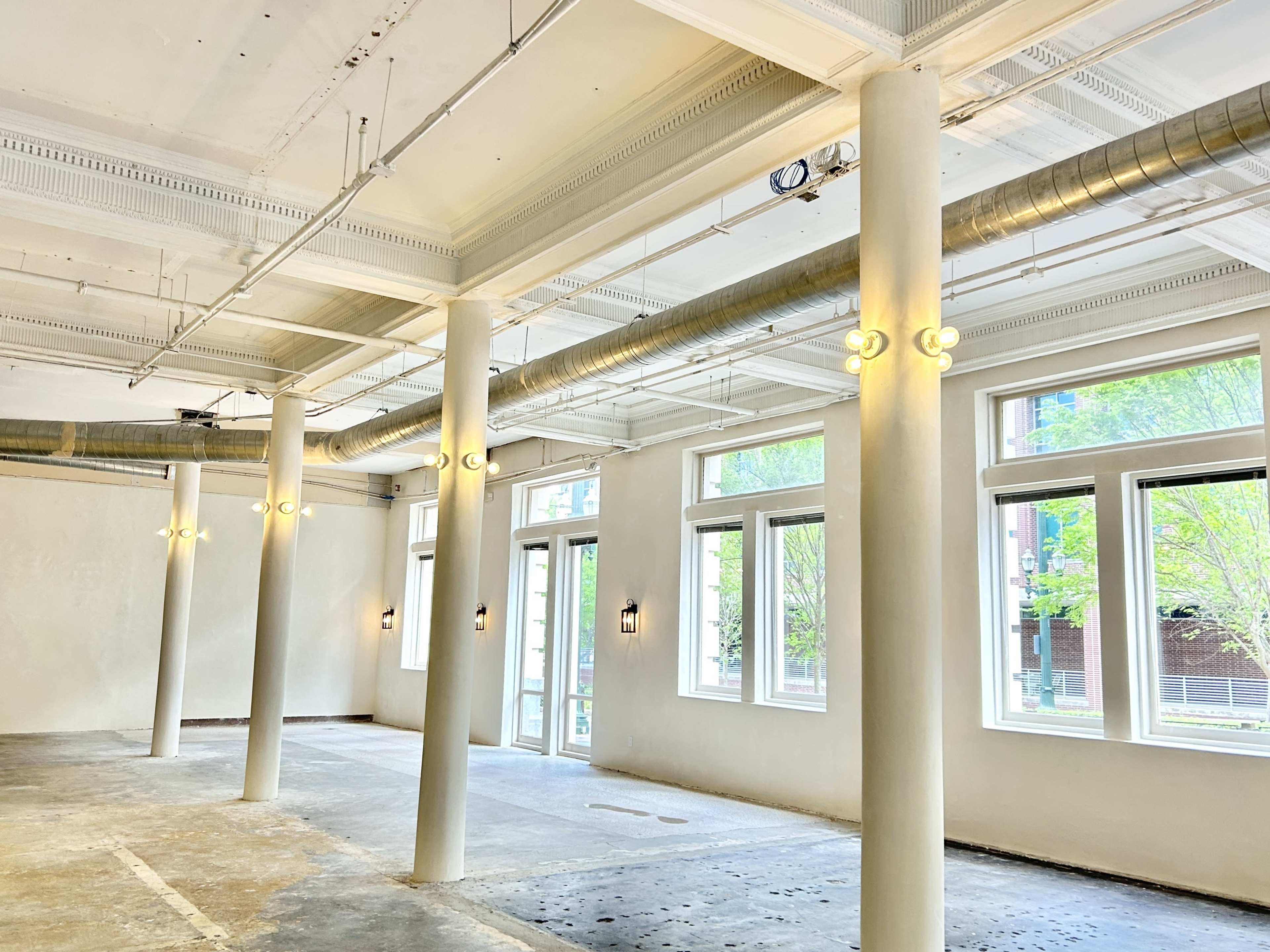 The image shows an empty interior space with white walls, concrete flooring, and large windows, featuring several columns and visible light fixtures.