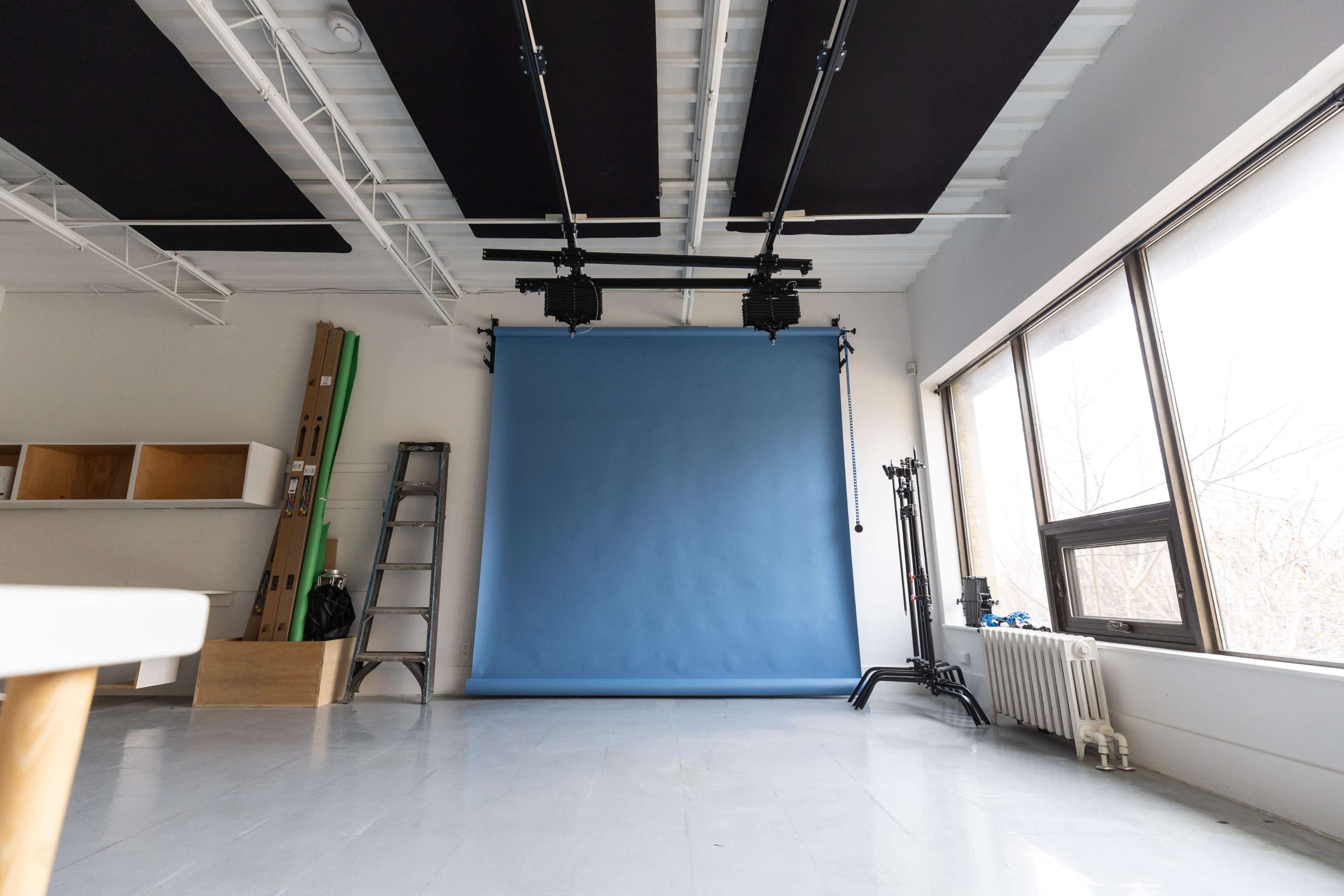 The image shows a photography studio with a blue backdrop, a ladder, lighting equipment, and large windows.