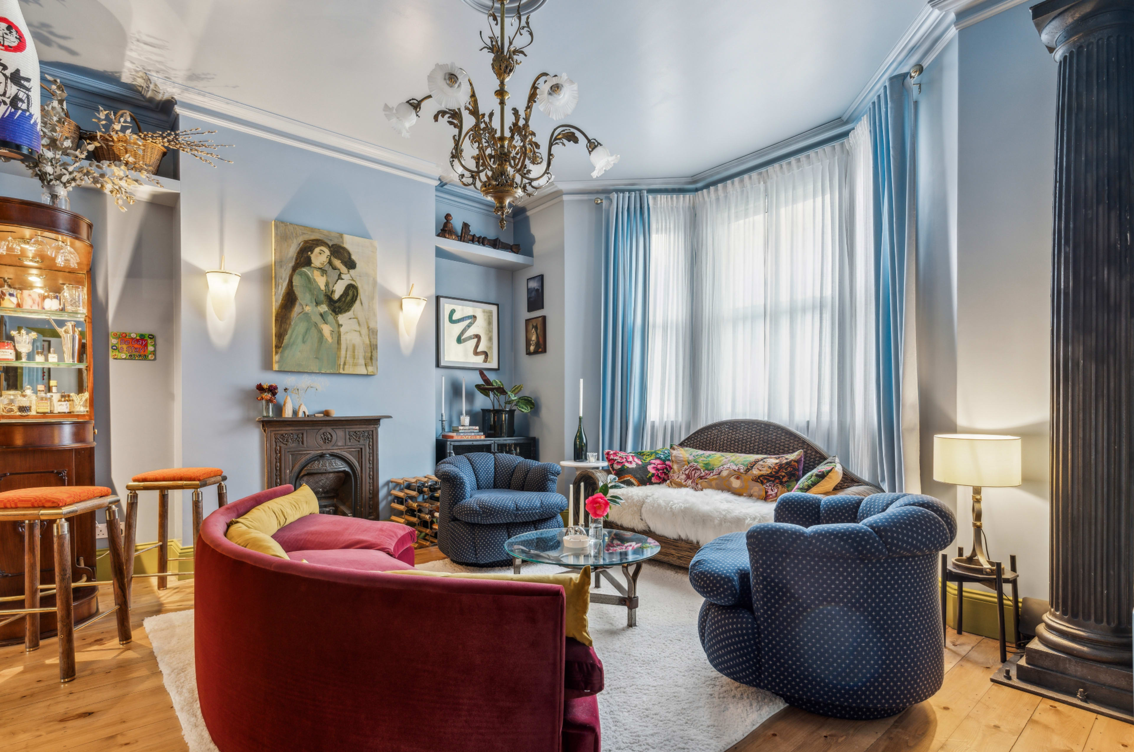 The living room features a mix of colorful seating, a decorative chandelier, and large windows that illuminate the space.
