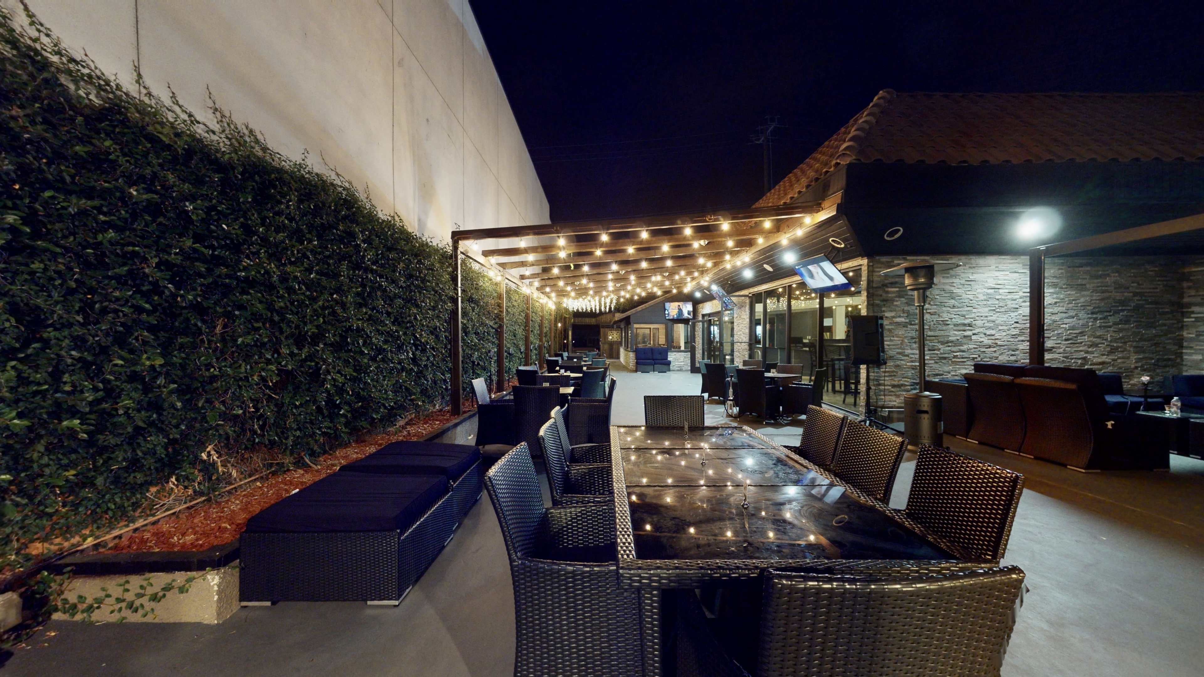 The image shows an outdoor seating area with wicker furniture and string lights overhead, adjacent to a wall of greenery, under a night sky.