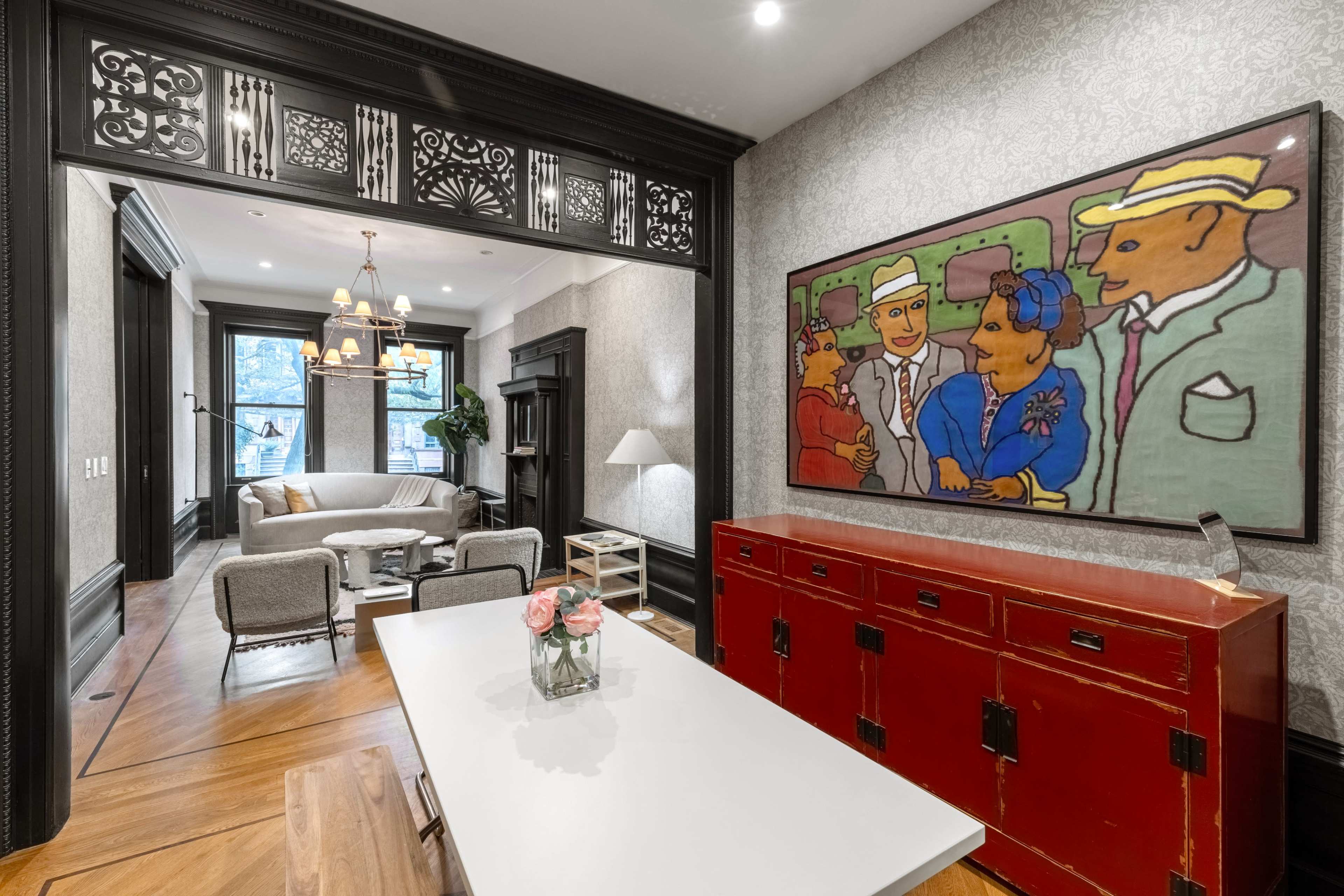 A modern interior features a dining area with a white table, a colorful artwork on the wall, and a red chest against a patterned background, leading into a living space with seating and large windows.