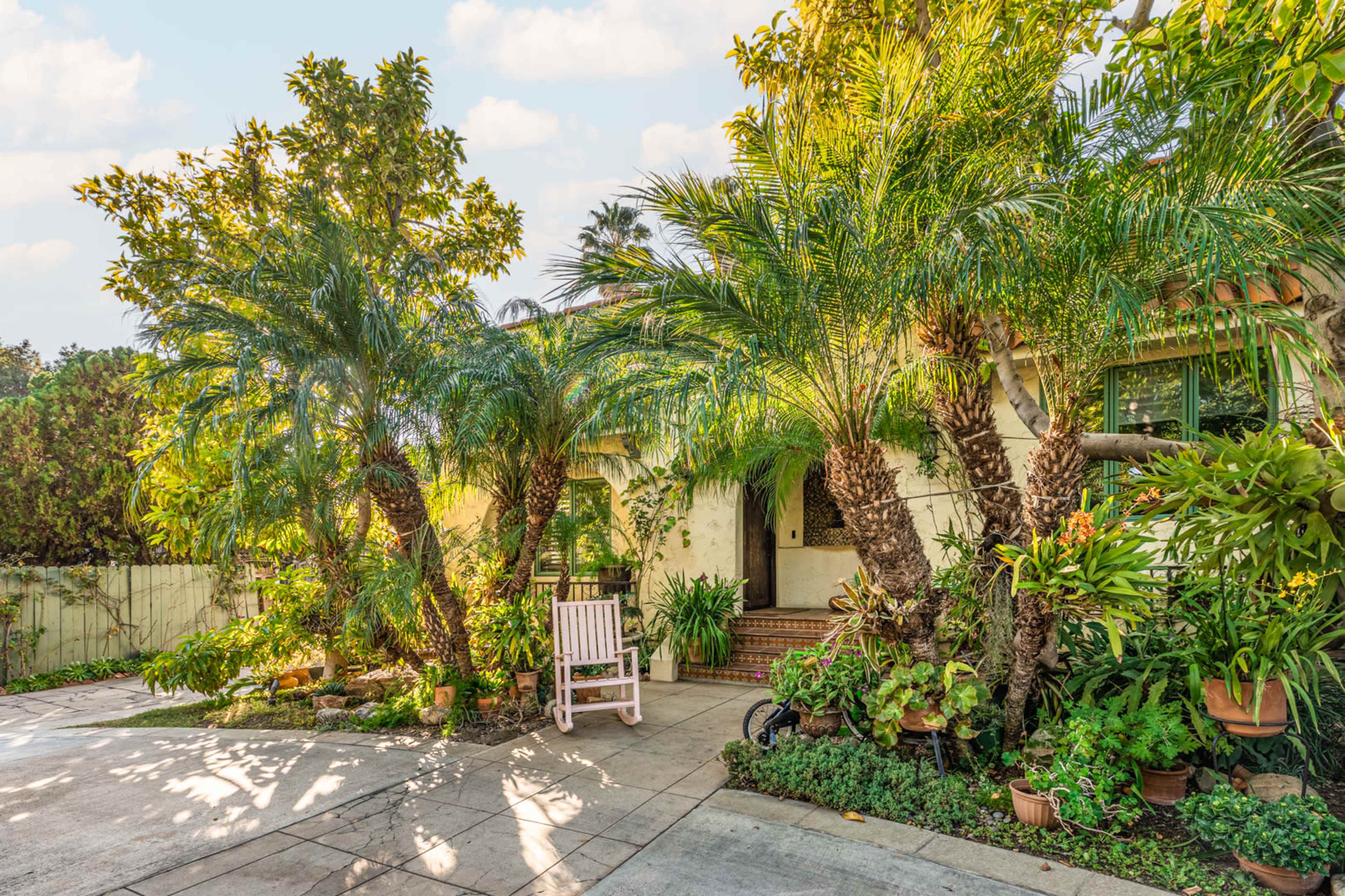 A front yard features a path leading to a house, surrounded by various tropical plants and a white rocking chair.