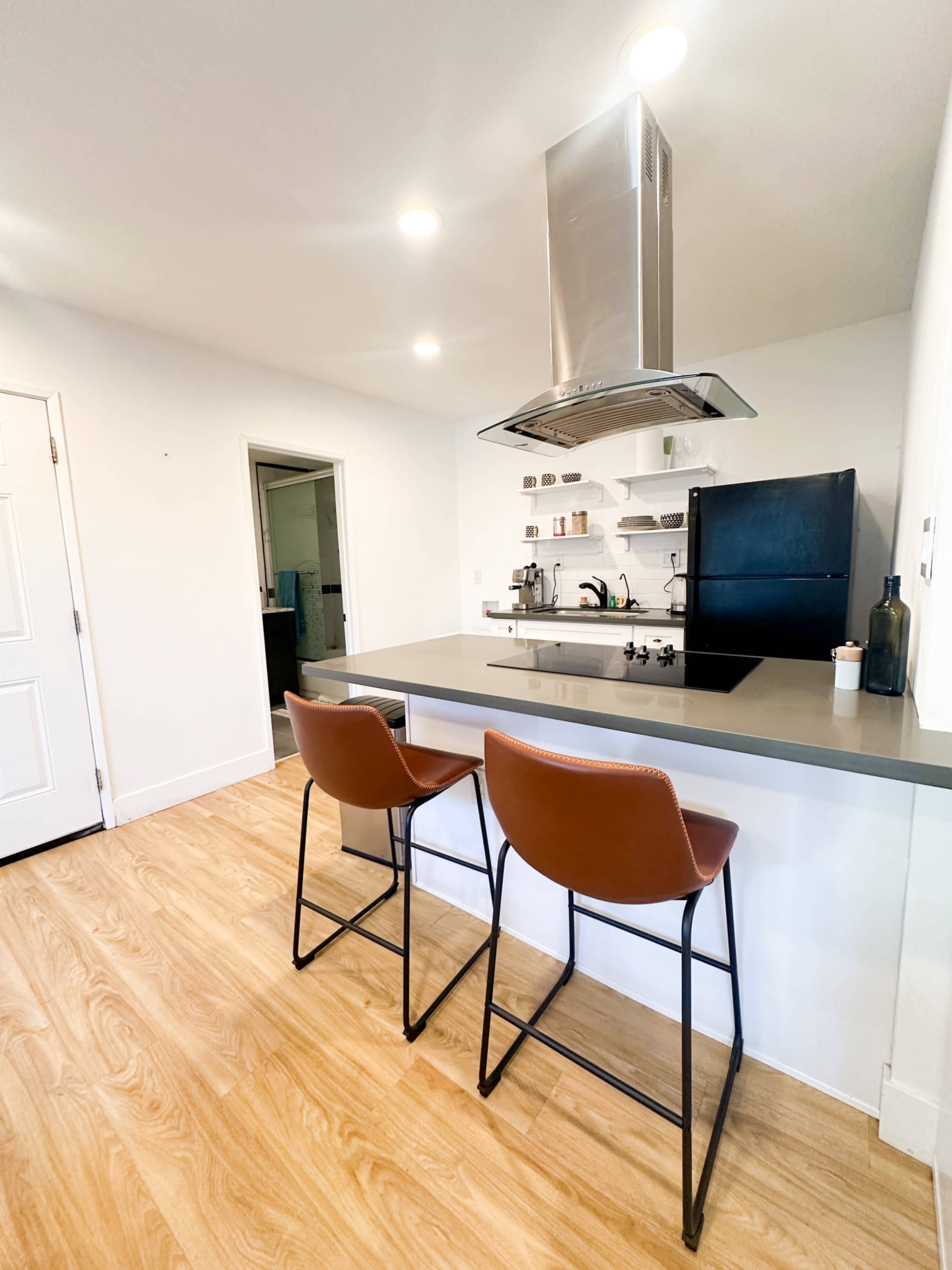 A modern kitchen features a central island with two leather bar stools, a black refrigerator, and a stainless steel range hood.