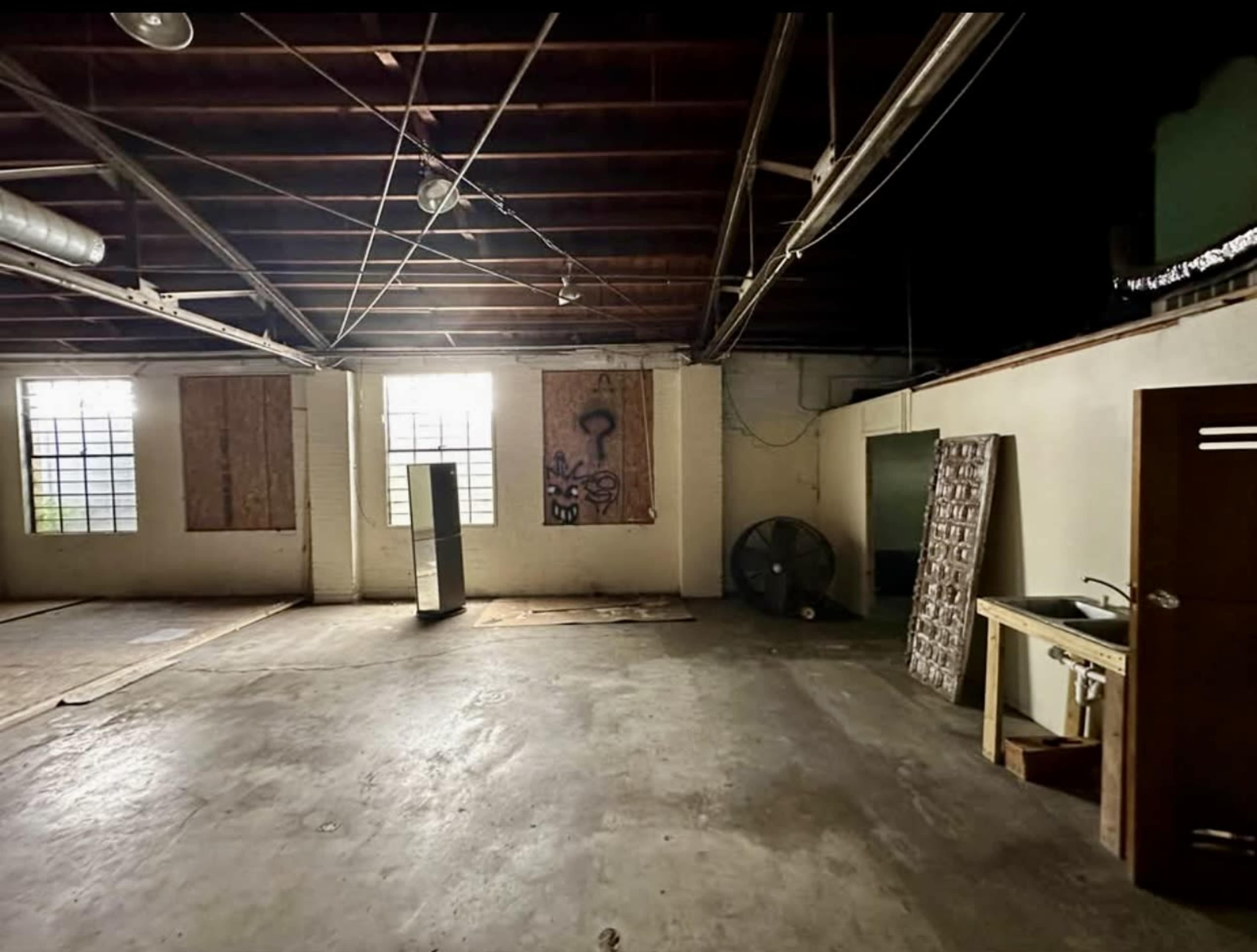 The image shows a large, empty indoor space with exposed beams, concrete floors, and boarded-up windows.