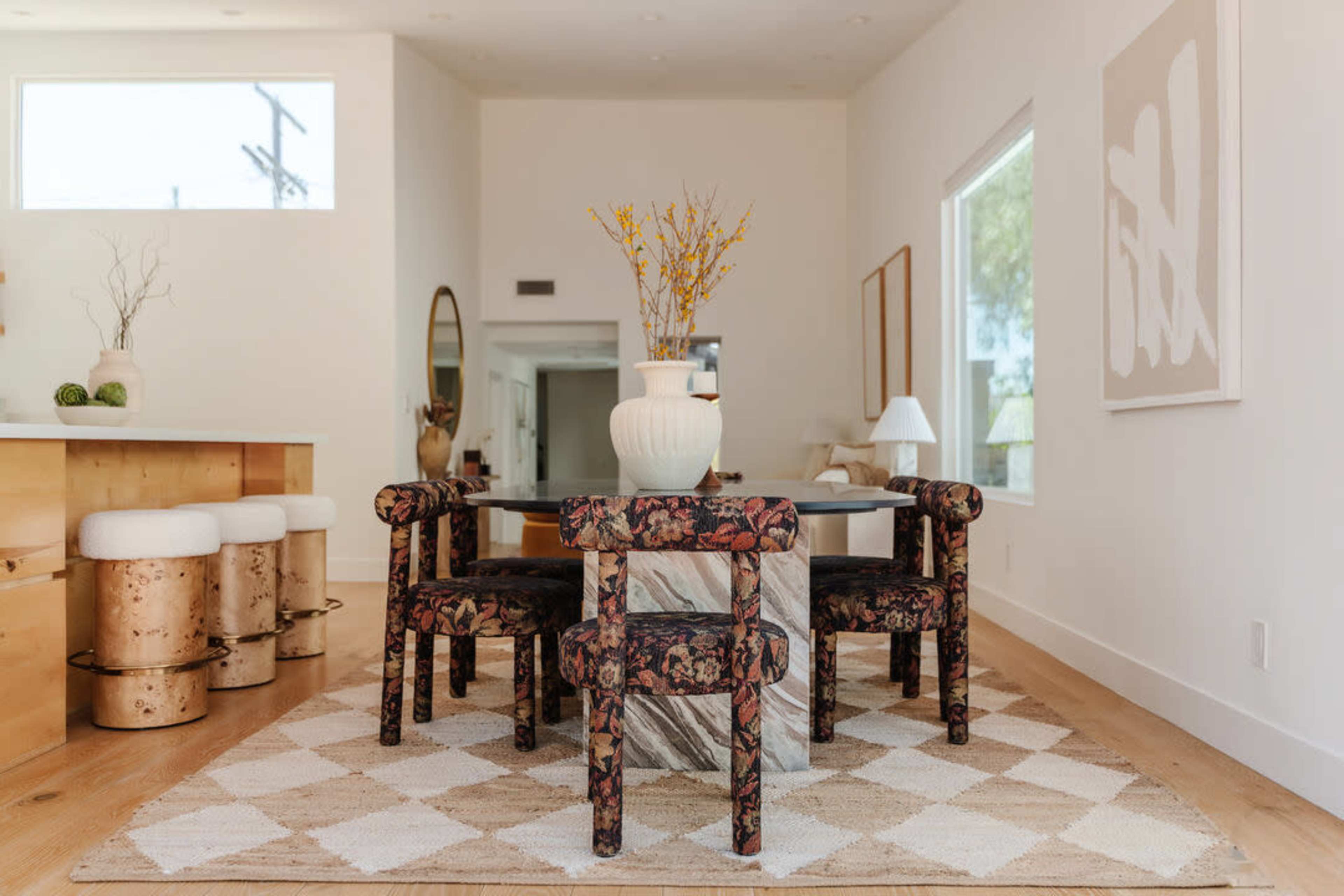A dining area features a round black table surrounded by upholstered chairs with floral patterns, set on a geometric rug.