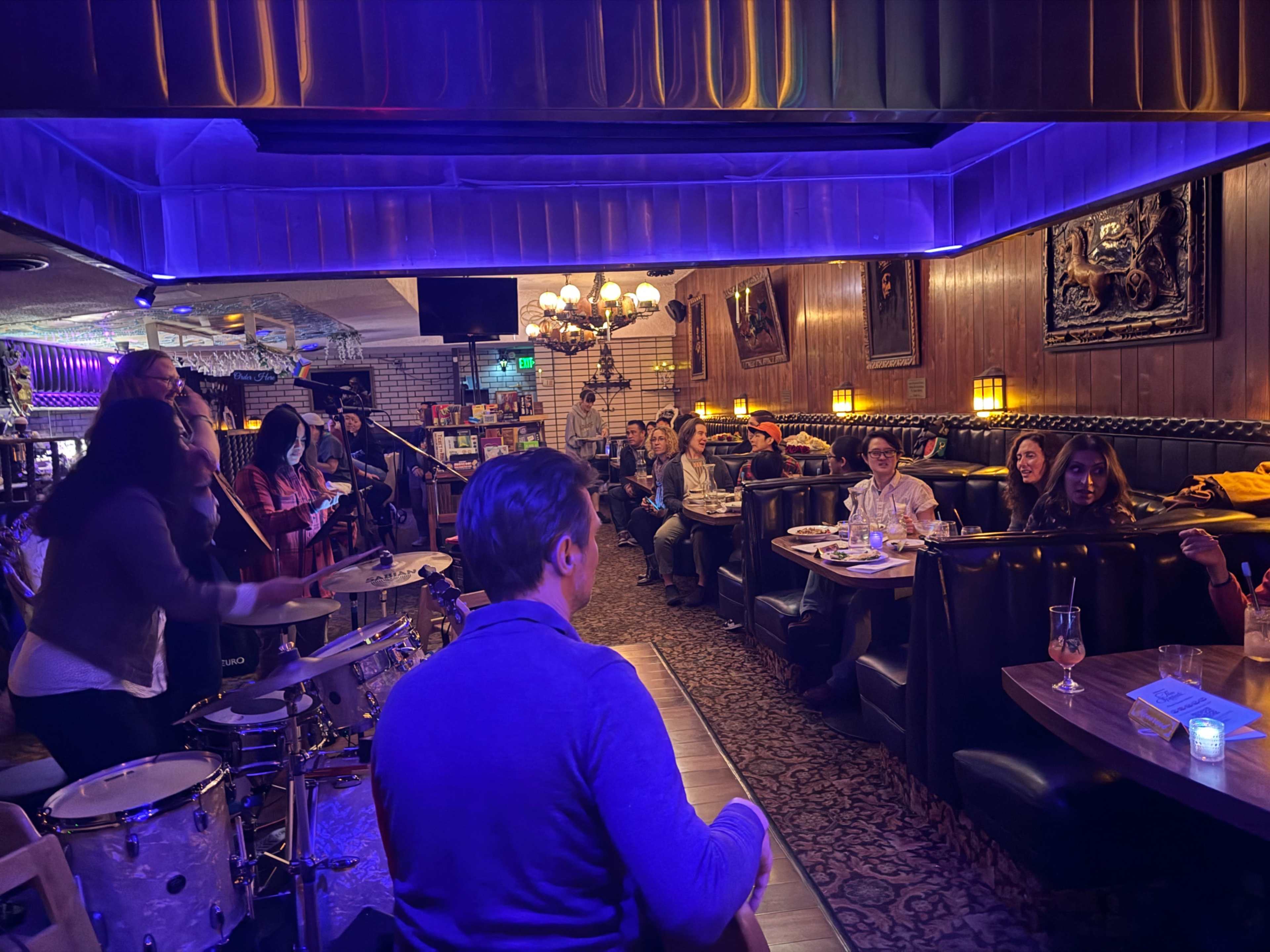 A live band performs on a small stage in a dimly lit bar filled with patrons seated at booths.