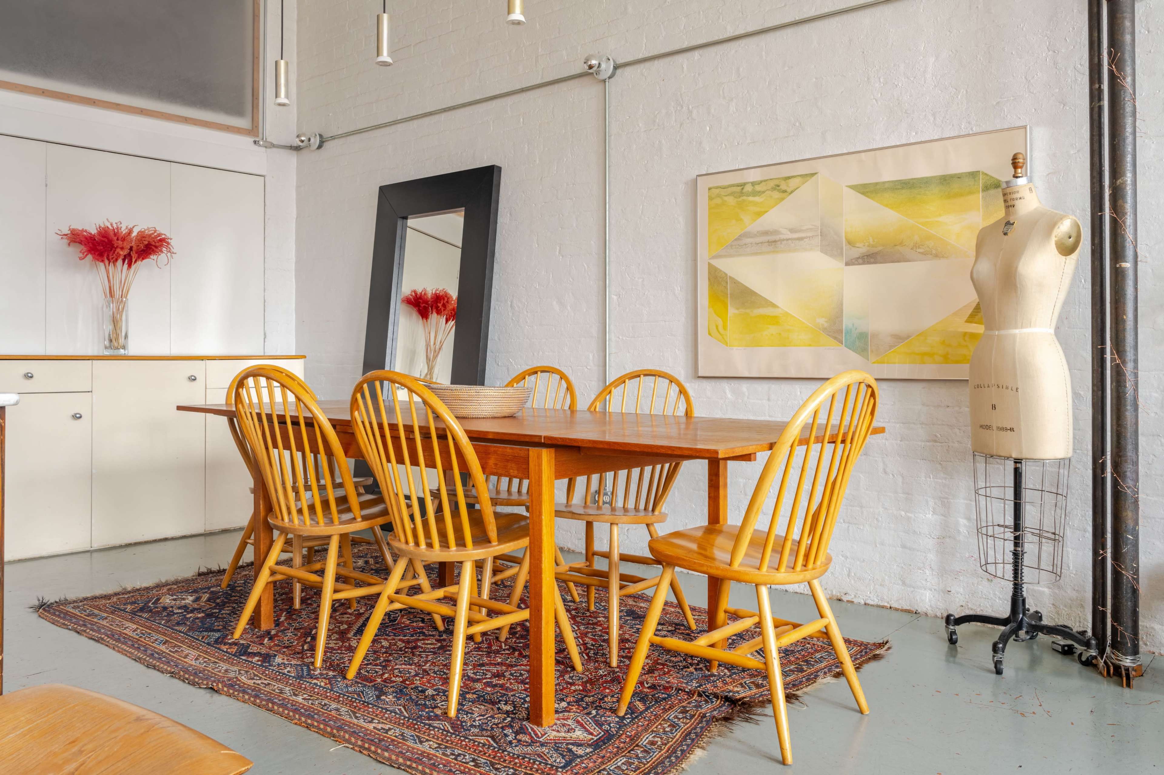 The image shows a dining area with a wooden table surrounded by six chairs, a large mirror on the wall, a decorative artwork, and an antique dress form in the corner.