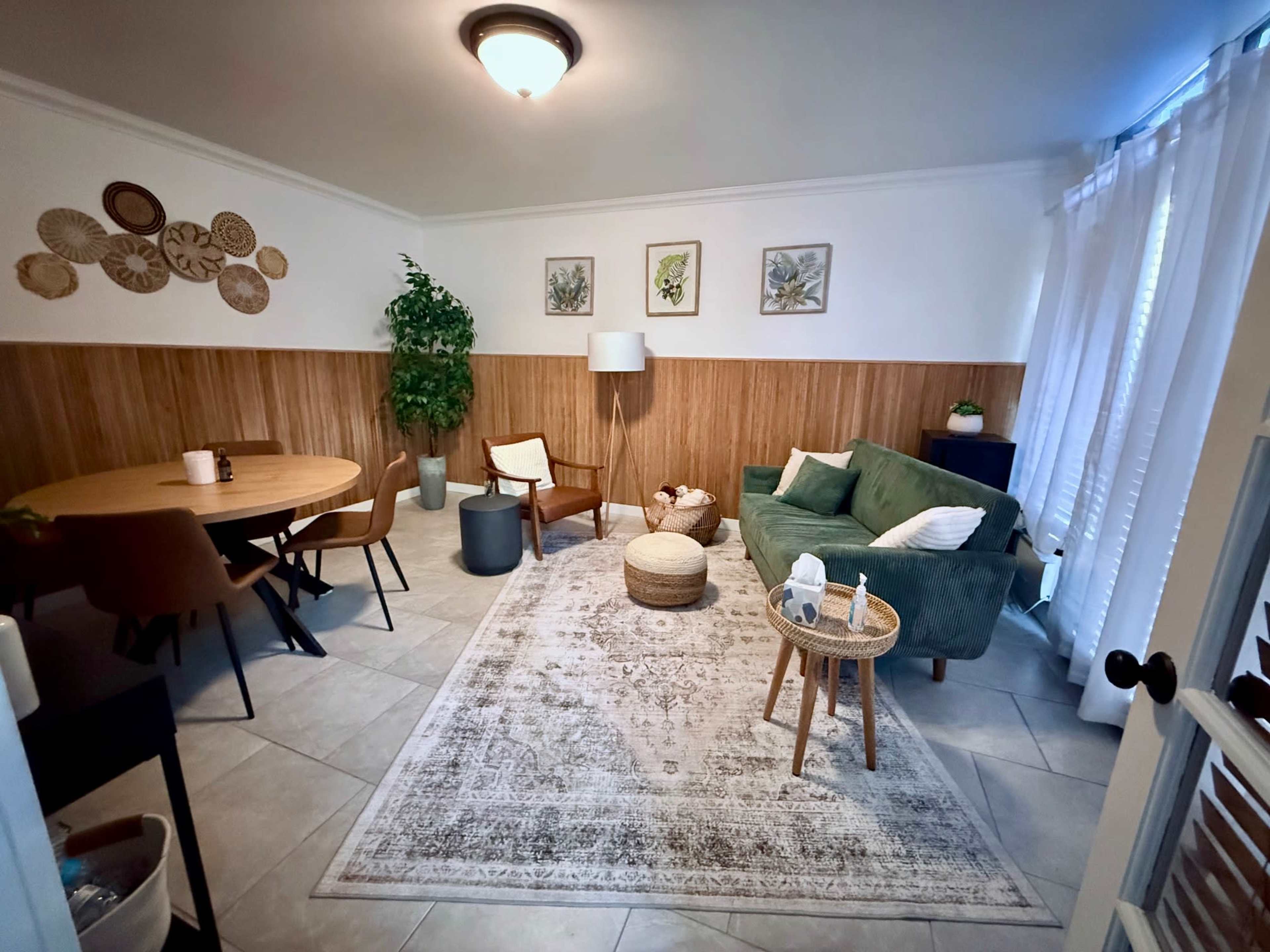 The image shows a cozy living area featuring a round dining table, a green sofa, a circular pouf, and decorative wall art.