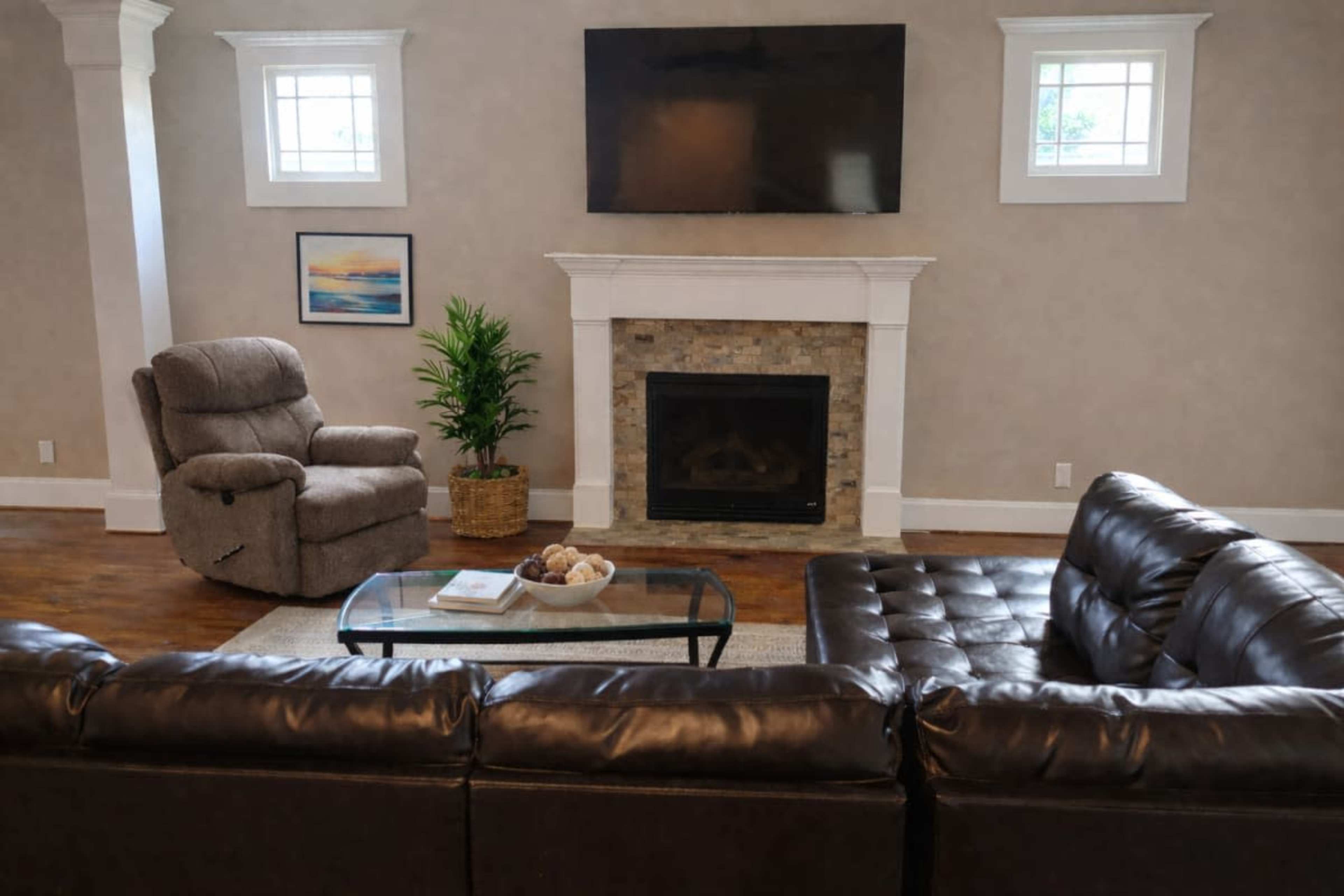 The living room features a leather sectional sofa, a glass coffee table, a recliner chair, a stone fireplace, and a flat-screen TV mounted on the wall.