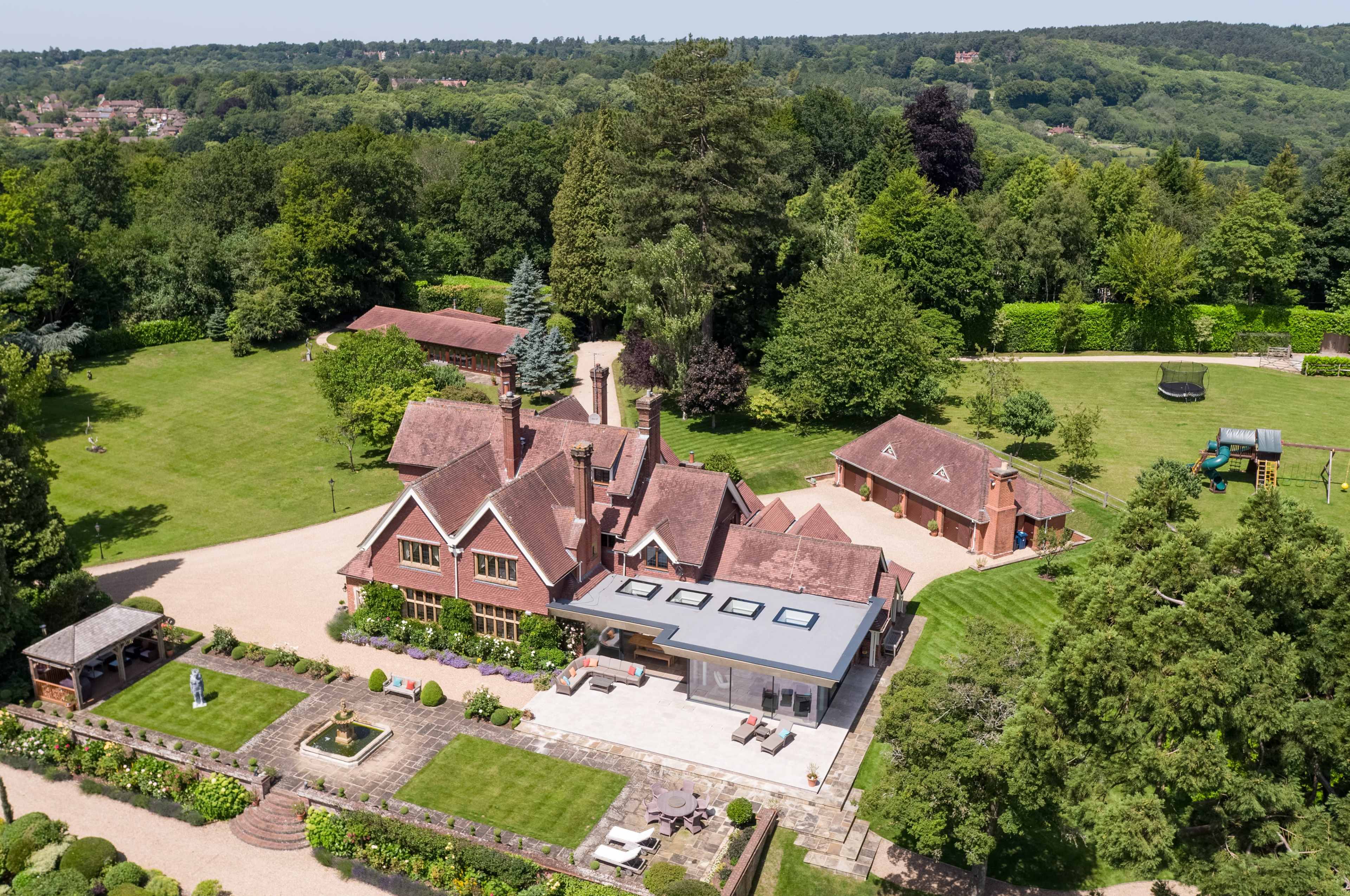 The image shows a large, red-brick house surrounded by well-maintained lawns and trees, featuring a modern extension with a flat roof and a landscaped garden area.
